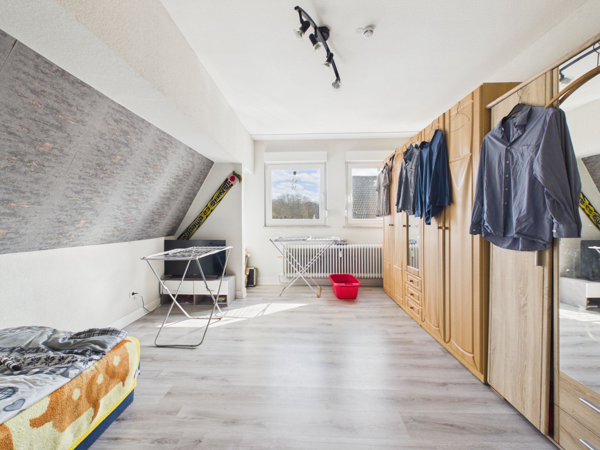 Helles Zimmer mit Kleiderschrank, Wäschegestellen, Bett und zwei Fenstern.