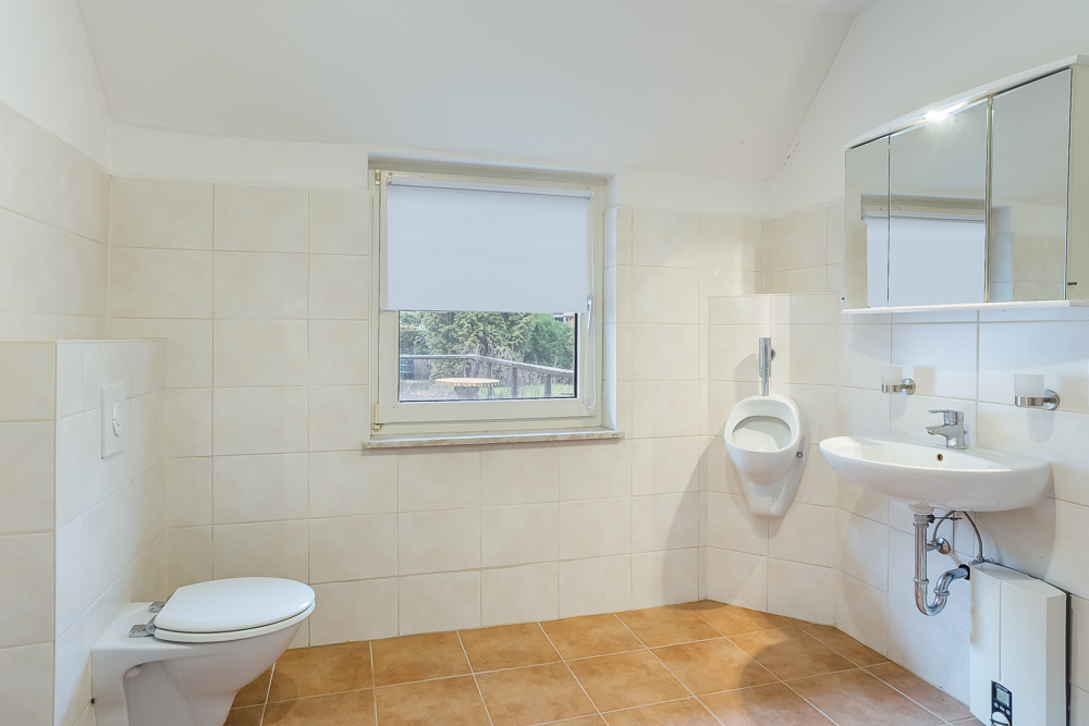 Helles Badezimmer mit WC, Urinal, Waschbecken und Spiegelschrank, Fenster mittig.