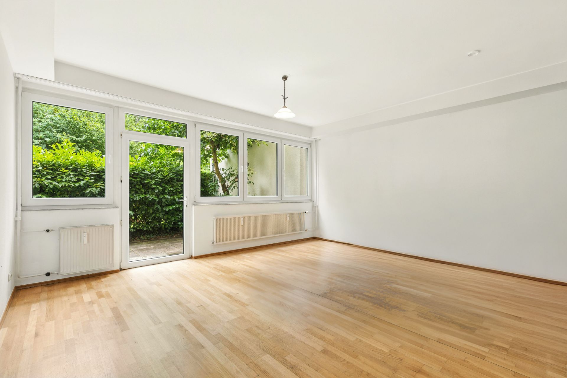 Heller, leerer Raum mit Holzfußboden, großen Fenstern und Tür zum Garten.