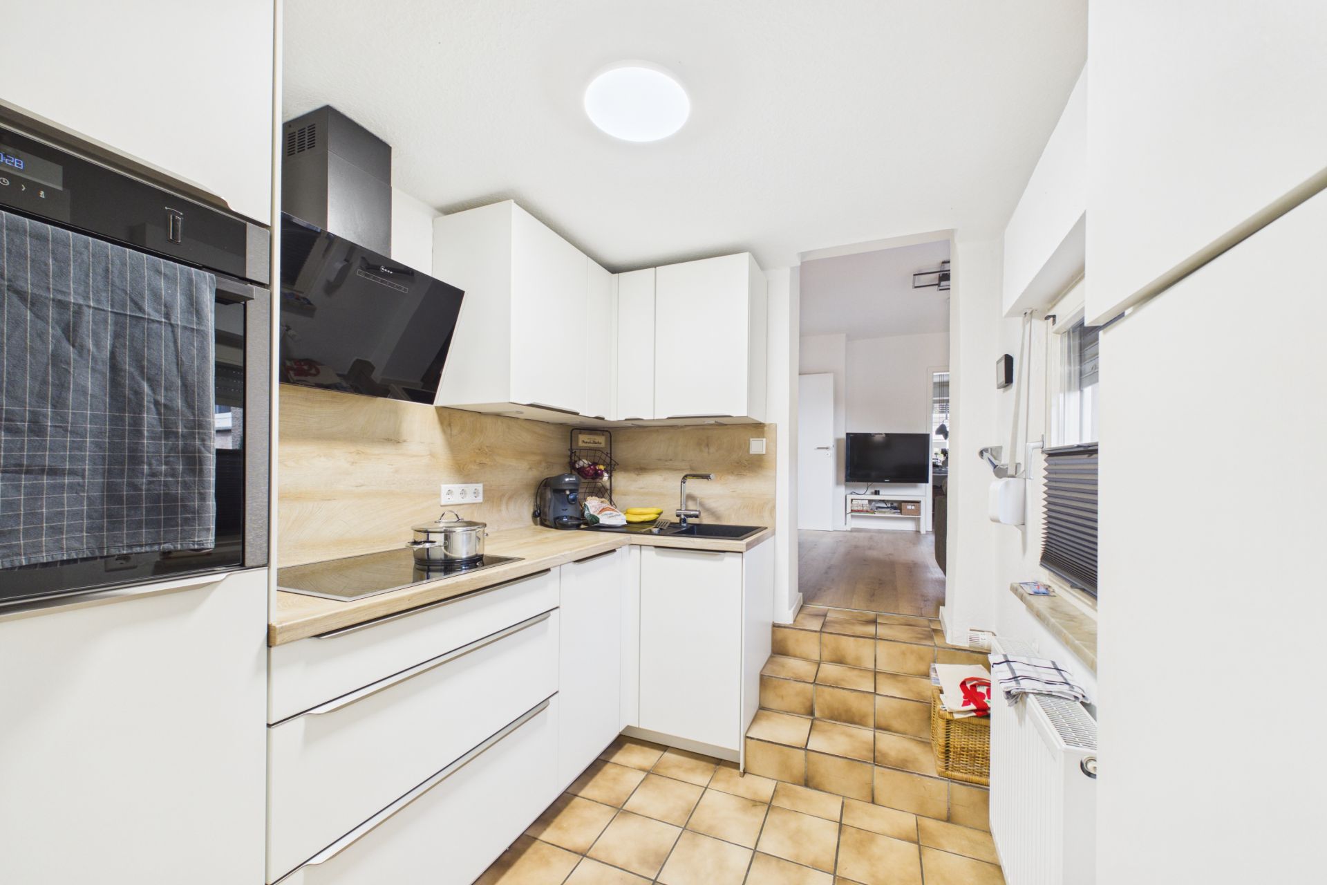 Moderne, weiße Küche mit Herd, Backofen und Fenster, Übergang zum Wohnzimmer im Hintergrund.