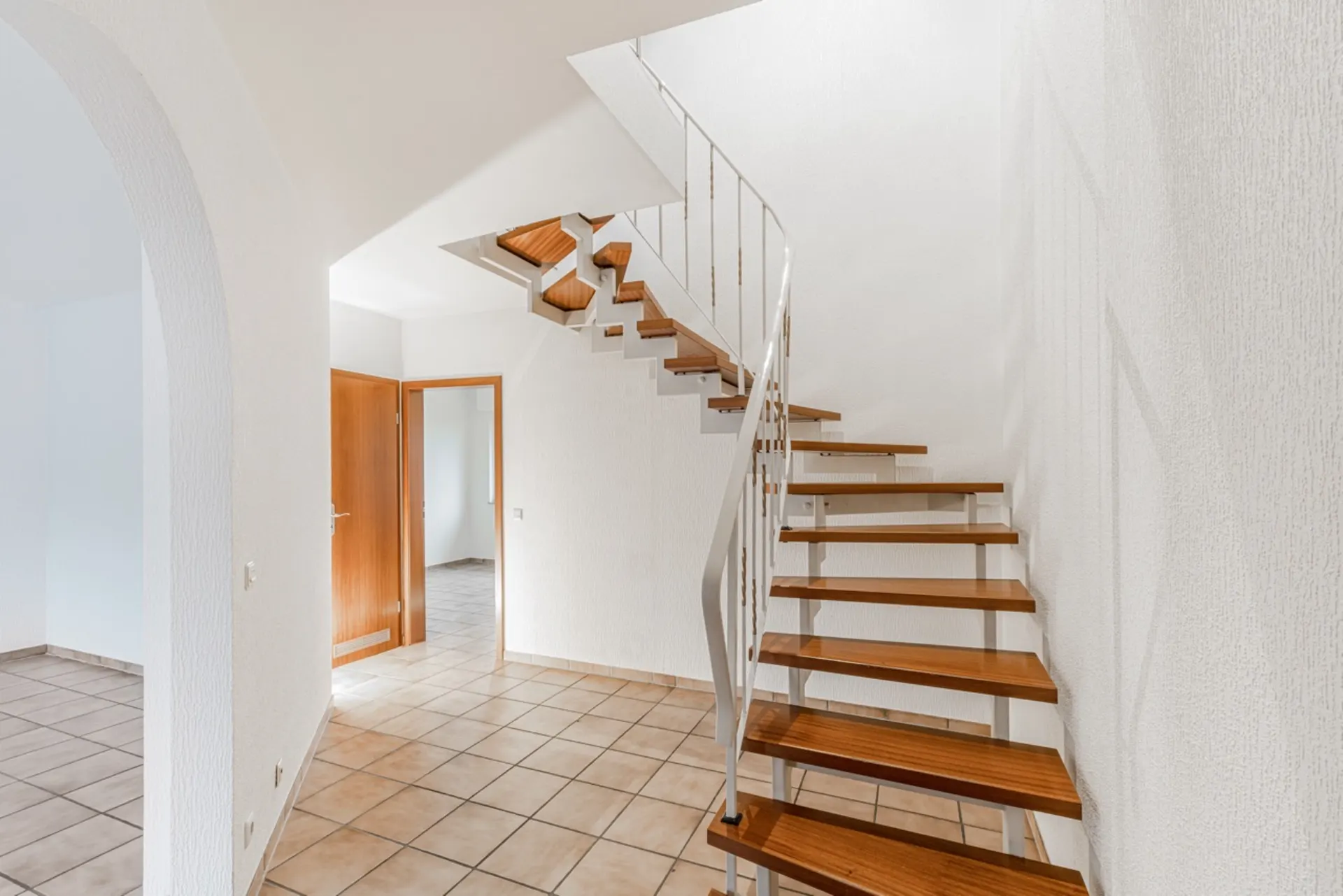 Heller Flur mit gefliesten Boden und moderner Holztreppe, weiße Wände, offene Tür zum Nebenraum.