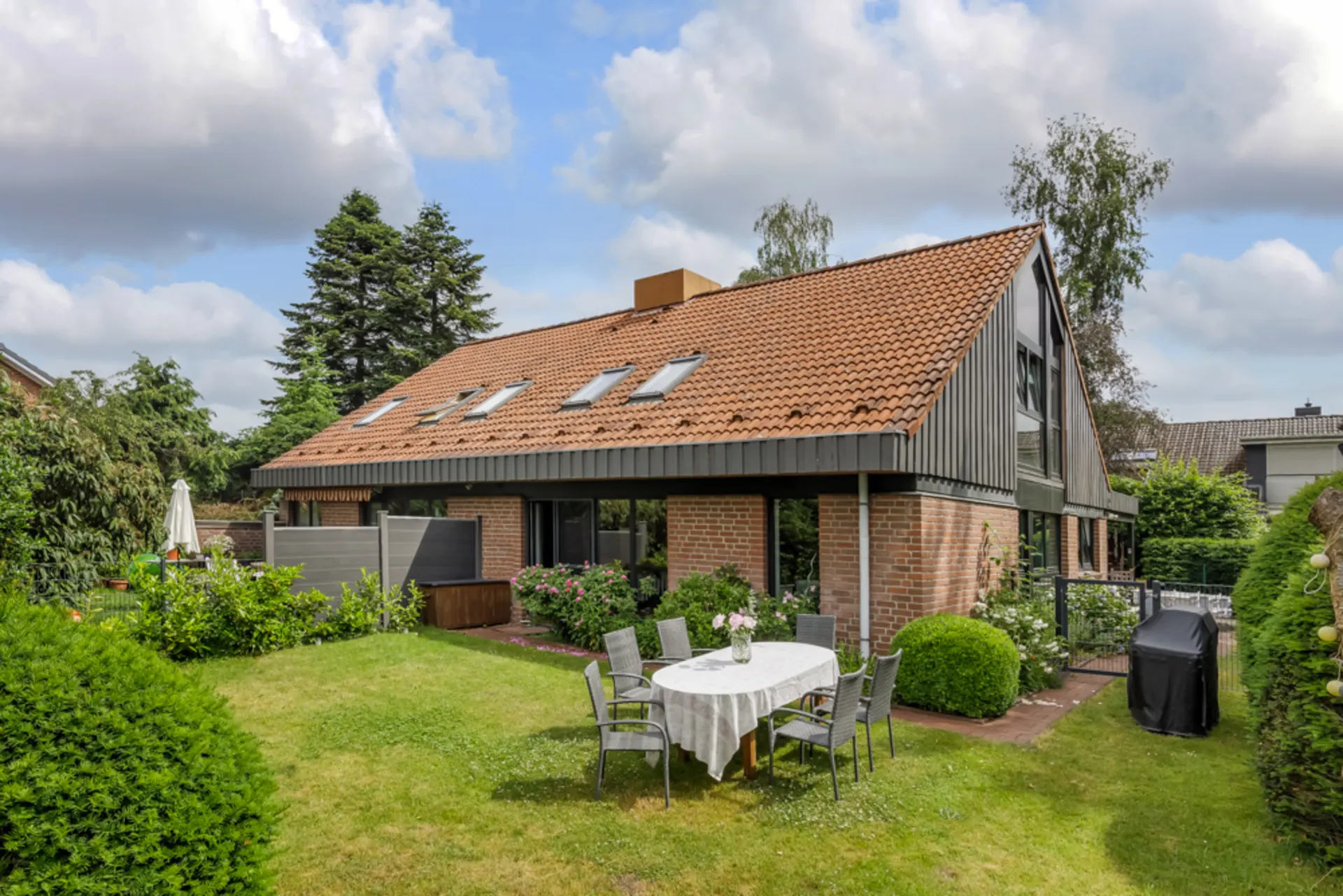 Backstein-Einfamilienhaus mit Satteldach, Garten, Terrassentisch und sechs Stühlen im Vordergrund.