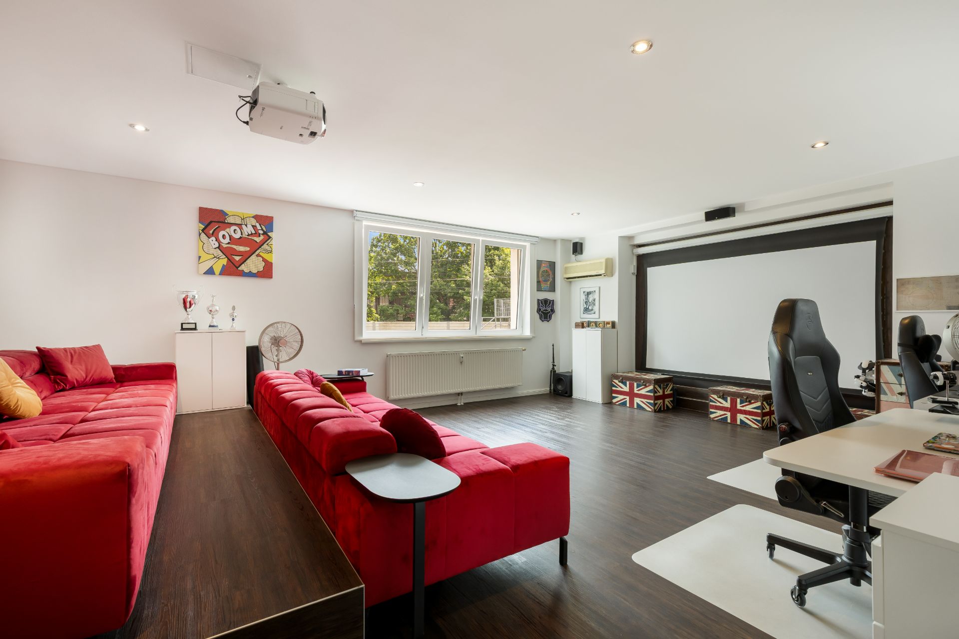 Helles Wohnzimmer mit zwei roten Sofas, großem Fenster, Schreibtisch und Leinwand.