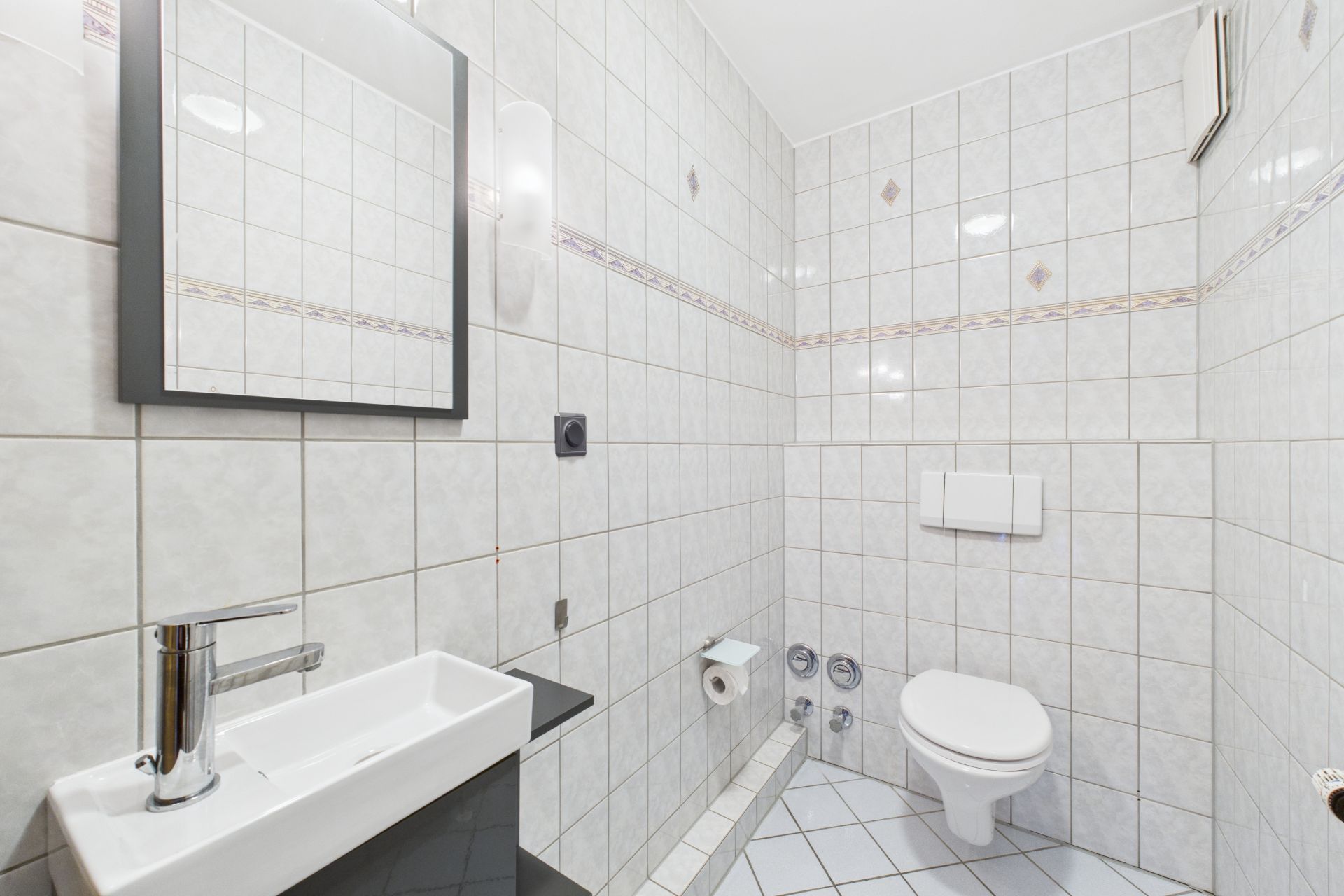 Modernes, weiß gefliestes Badezimmer mit Wandtoilette, Waschbecken und Spiegel.