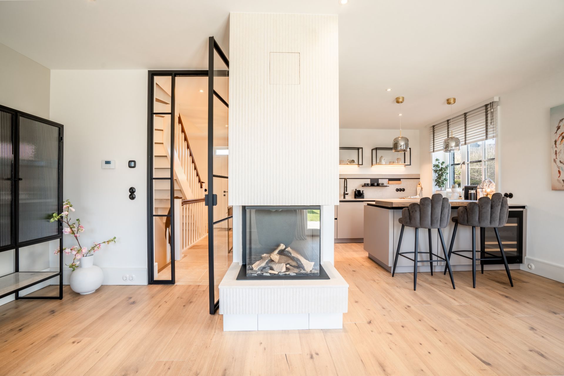 Helles Wohnzimmer mit zentralem Kamin, Holzboden und offener Küche mit Barhockern.