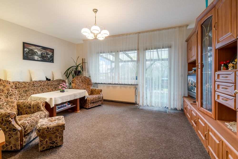 Wohnzimmer mit gemusterten Polstermöbeln, Wohnwand, Tisch und großen Fenstern mit weißen Vorhängen.
