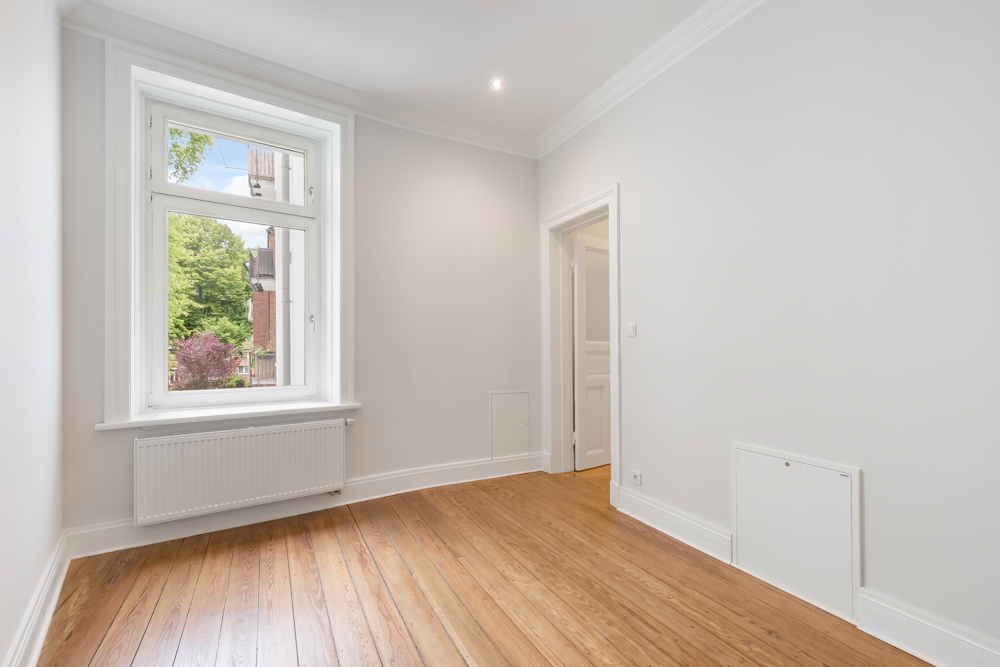 Helles, leeres Zimmer mit Holzfußboden, weißer Wand und großem Fenster.