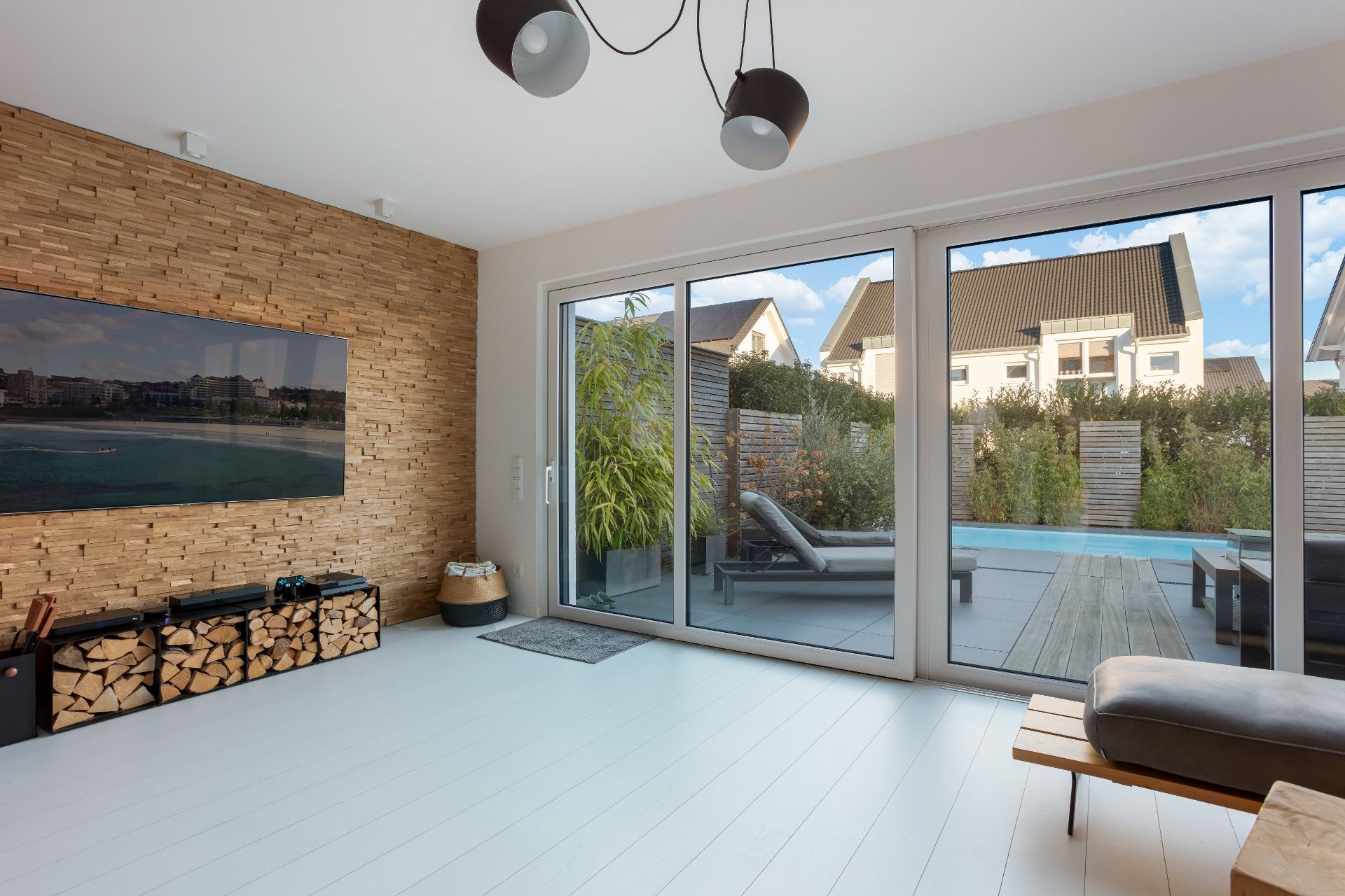 Modernes Wohnzimmer mit Holzwand, Kaminholz, großer Glasfront und Blick auf Terrasse und Pool.