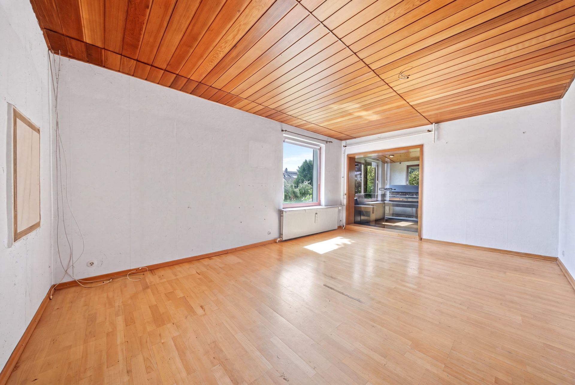 Leerer Raum mit Holzdecke und -boden, großem Fenster und Zugang zu einer angrenzenden Küche.