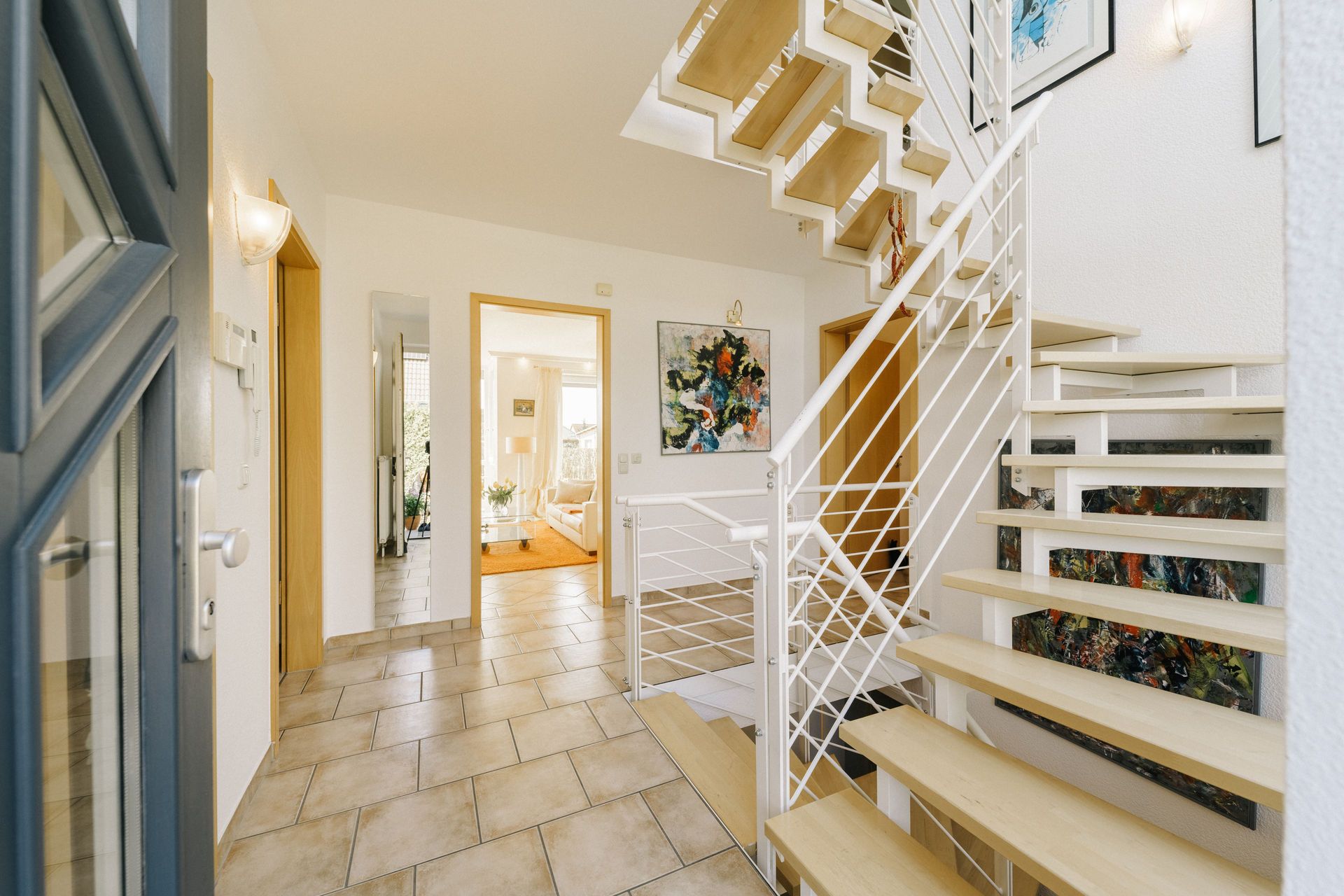 Heller Flur mit moderner Holztreppe, Fliesenboden und Blick in ein Wohnzimmer.