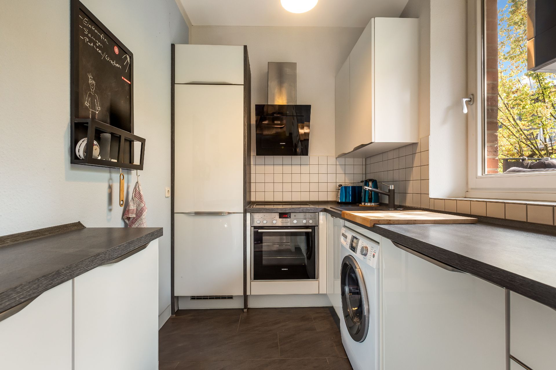 Moderne Küche mit weißen Schränken, Herd, Waschmaschine und Fenster mit Blick nach draußen.