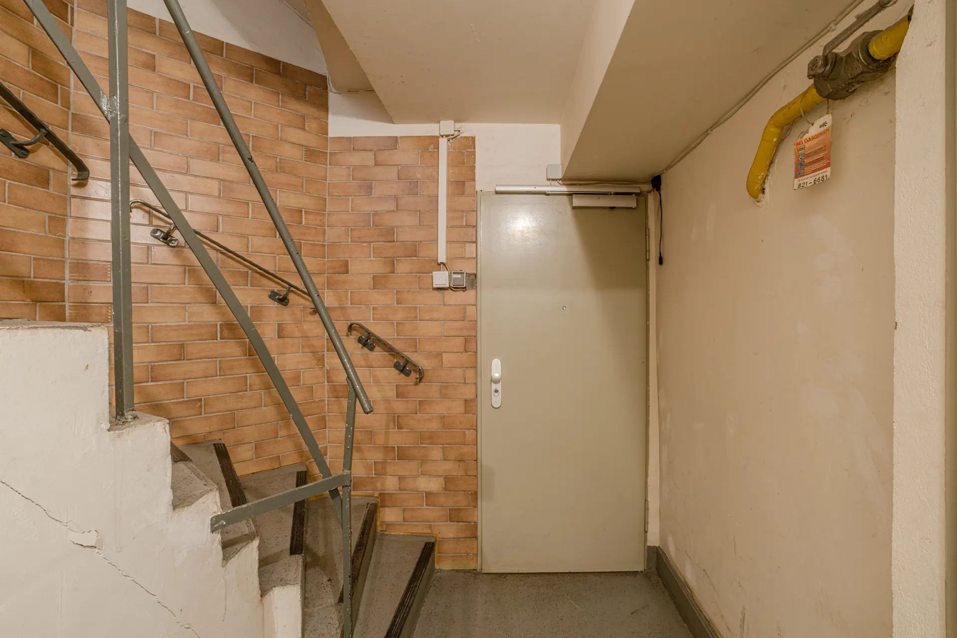 Treppenhaus mit braunen Fliesen, Geländer und grauer Tür neben gelbem Rohr.