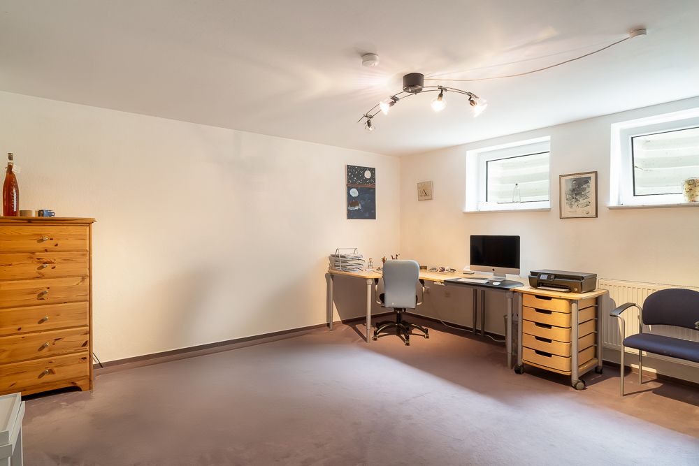 Helles Arbeitszimmer mit Schreibtisch, Computer, Holzkommode und zwei Fenstern.