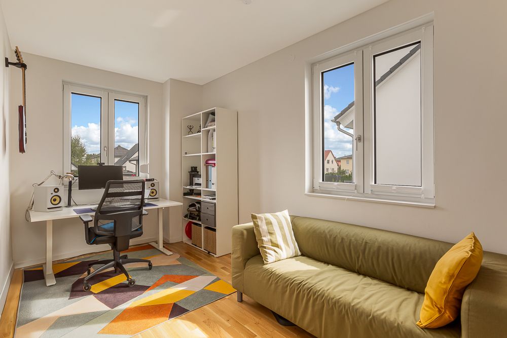 Helles Arbeitszimmer mit Schreibtisch, Regal, grünem Sofa und Teppich mit buntem Muster.