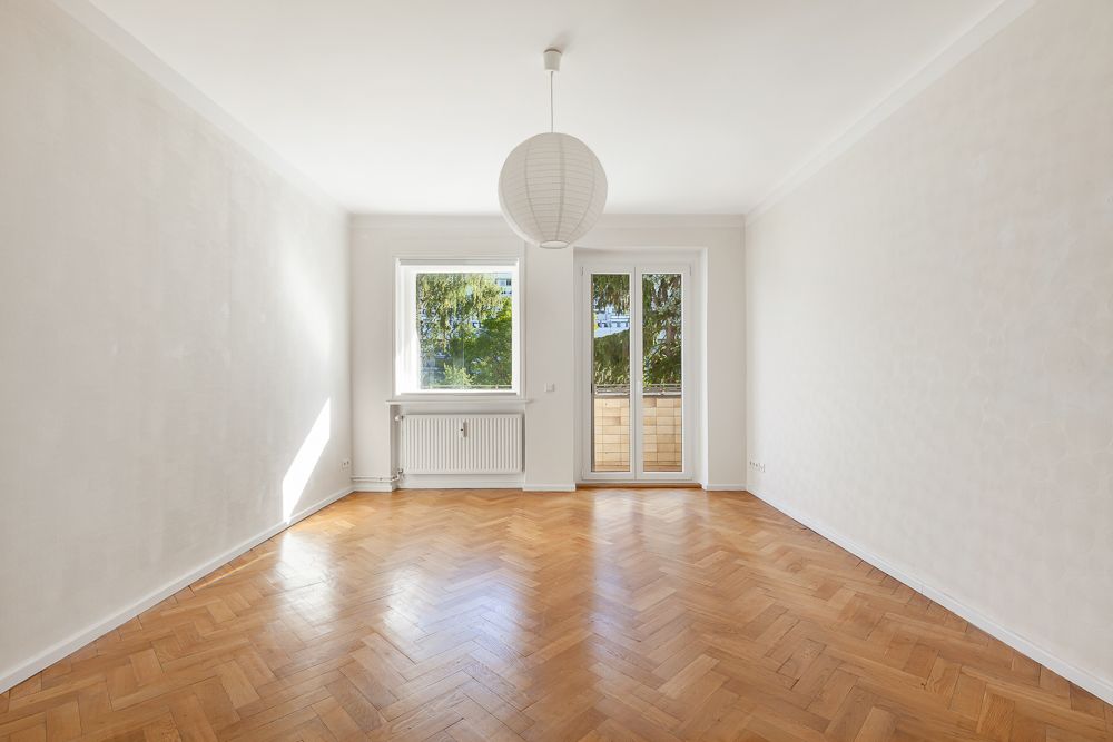 Helles, leeres Zimmer mit Parkettboden, Fenster, Balkontür und Papierlampe an der Decke.
