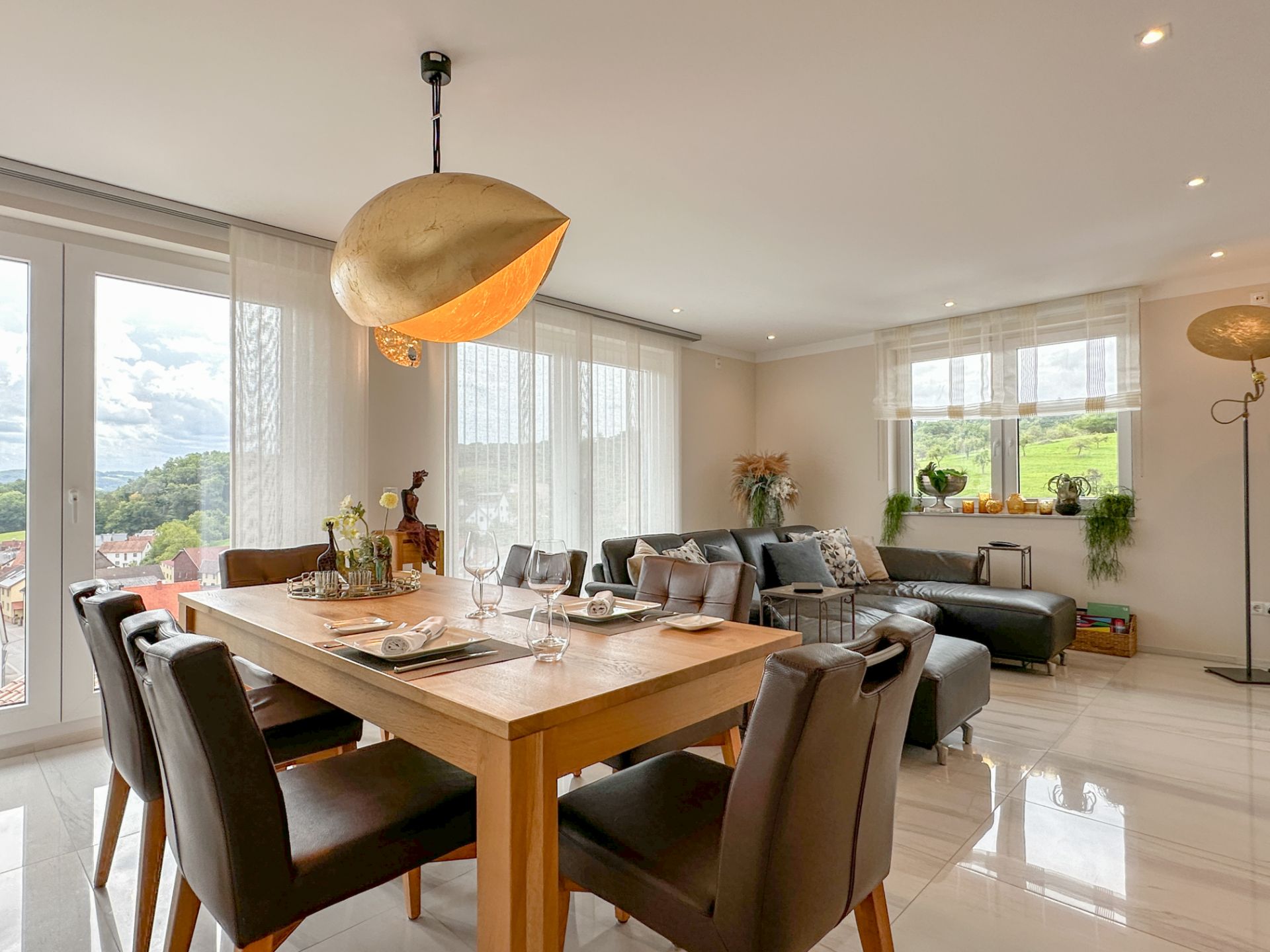 Helles Wohnzimmer mit Esstisch, sechs Stühlen, Sofa und großen Fenstern mit Blick ins Grüne.