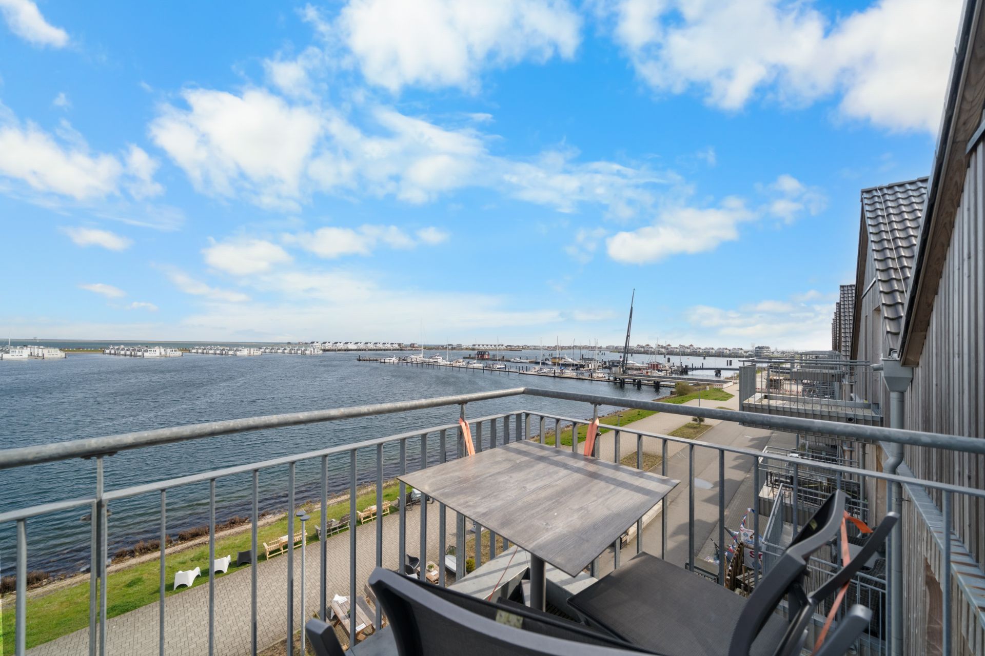 Balkon mit Tisch und Stühlen, Blick auf Wasser, Promenade und Yachthafen bei blauem Himmel.