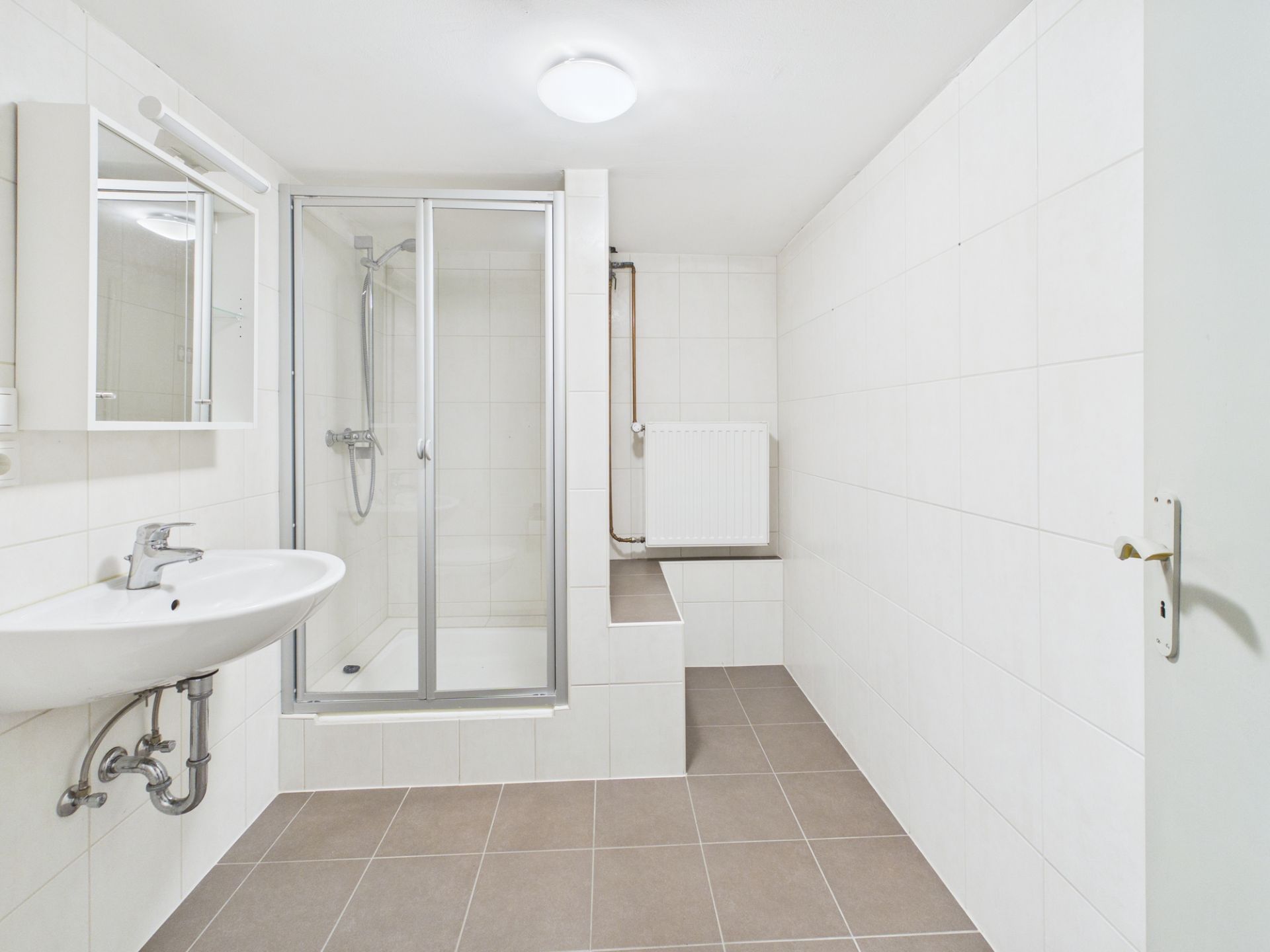 Helles Badezimmer mit Dusche, Waschbecken und Spiegel, weiße Fliesen und Heizkörper.