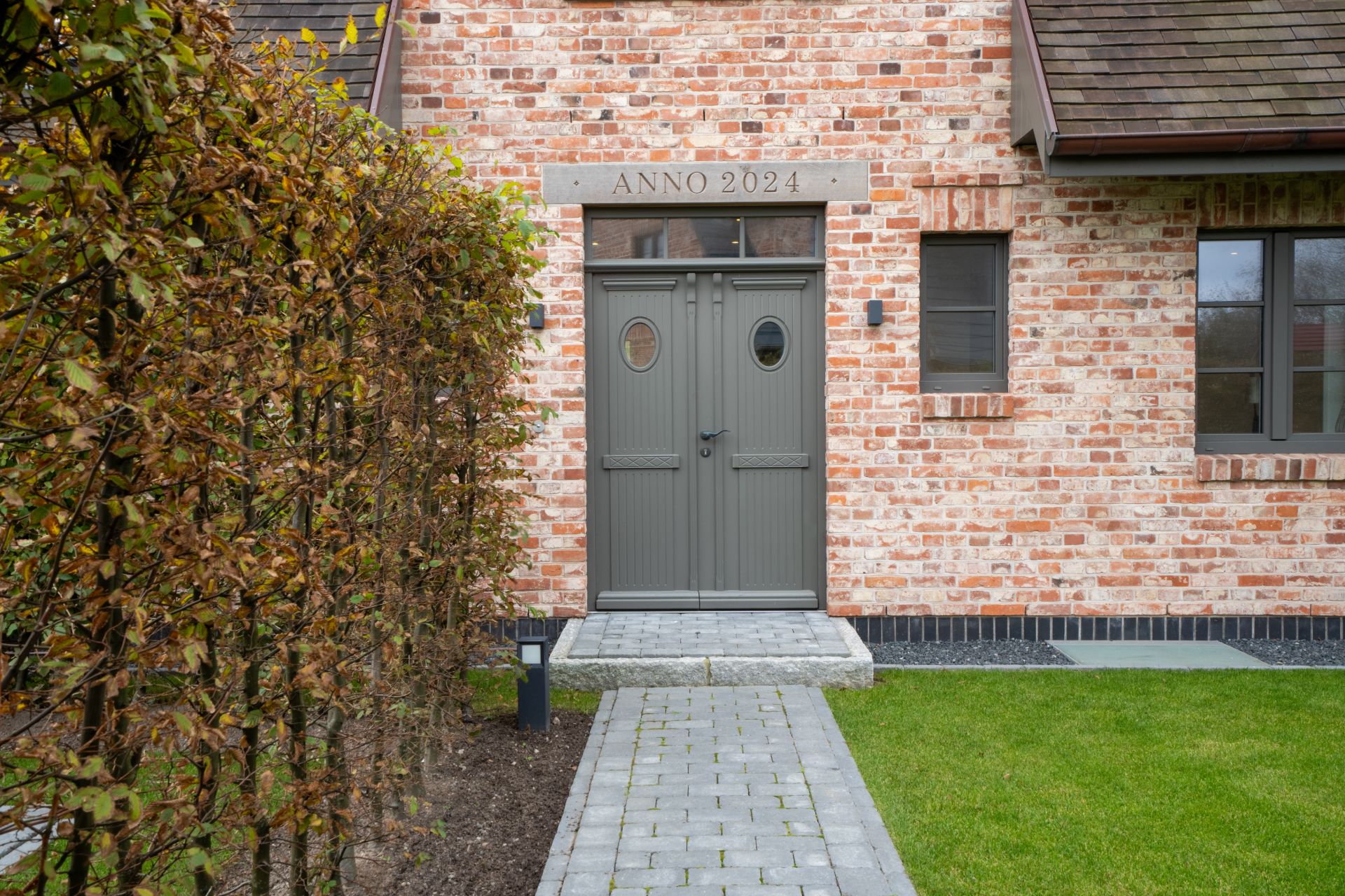 Backsteinfassade mit grauer Haustür, Gehweg und Hecke an der Seite.