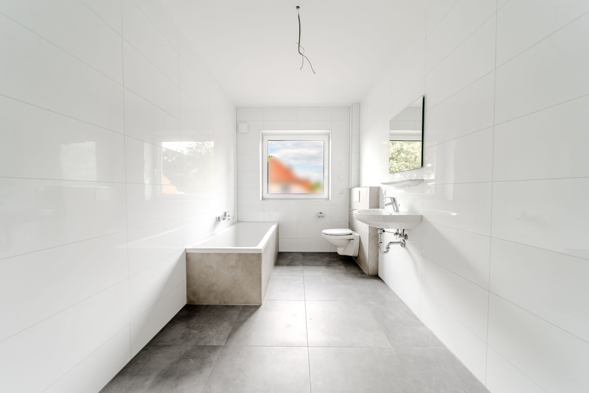 Modernes, weiß gefliestes Badezimmer mit Badewanne, Toilette, Waschbecken und großem Fenster.