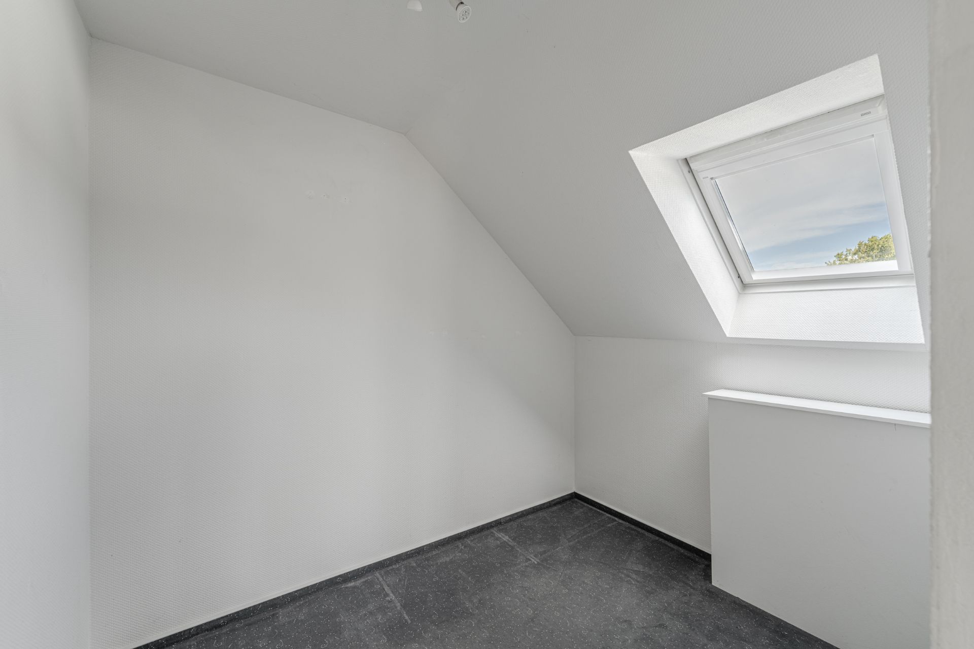 Kleiner, leerer Raum mit schräger Decke und Dachfenster, weiße Wände, dunkler Boden.
