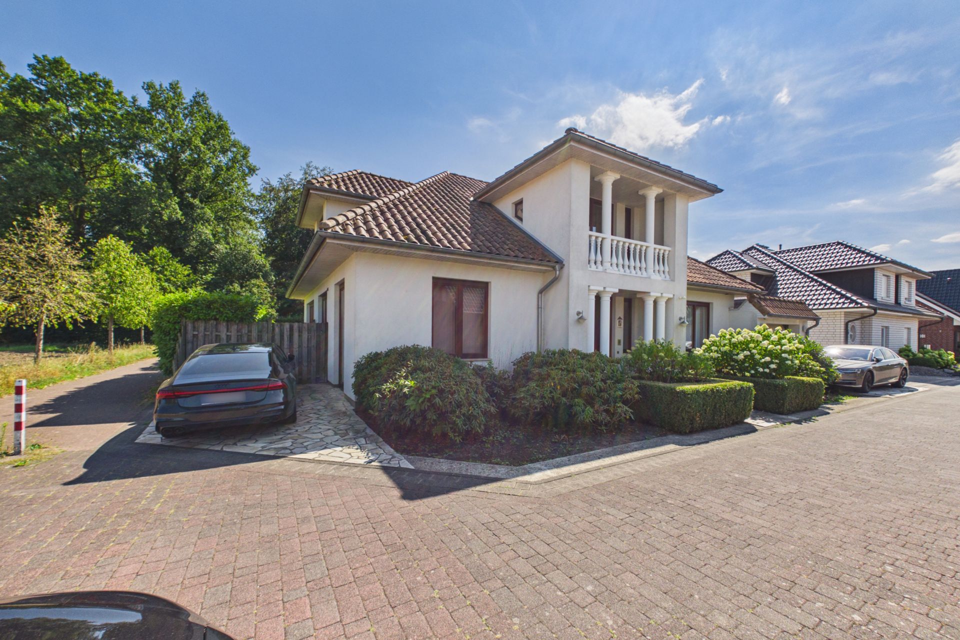 Weißes Einfamilienhaus mit Balkon, Garten und geparkten Autos auf der Einfahrt.