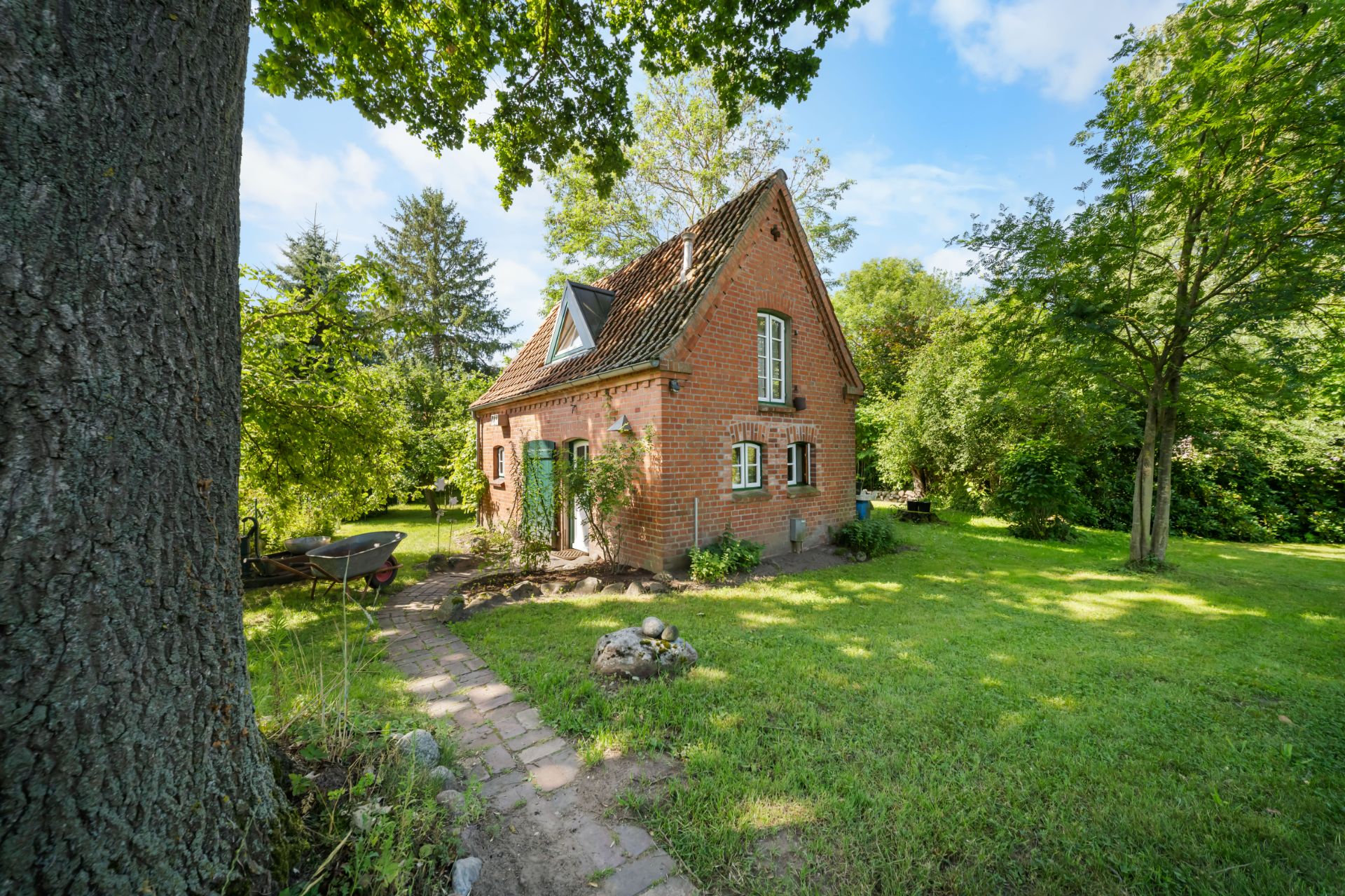Kleines Backsteinhaus mit Satteldach im Grünen, umgeben von Bäumen und Rasen.