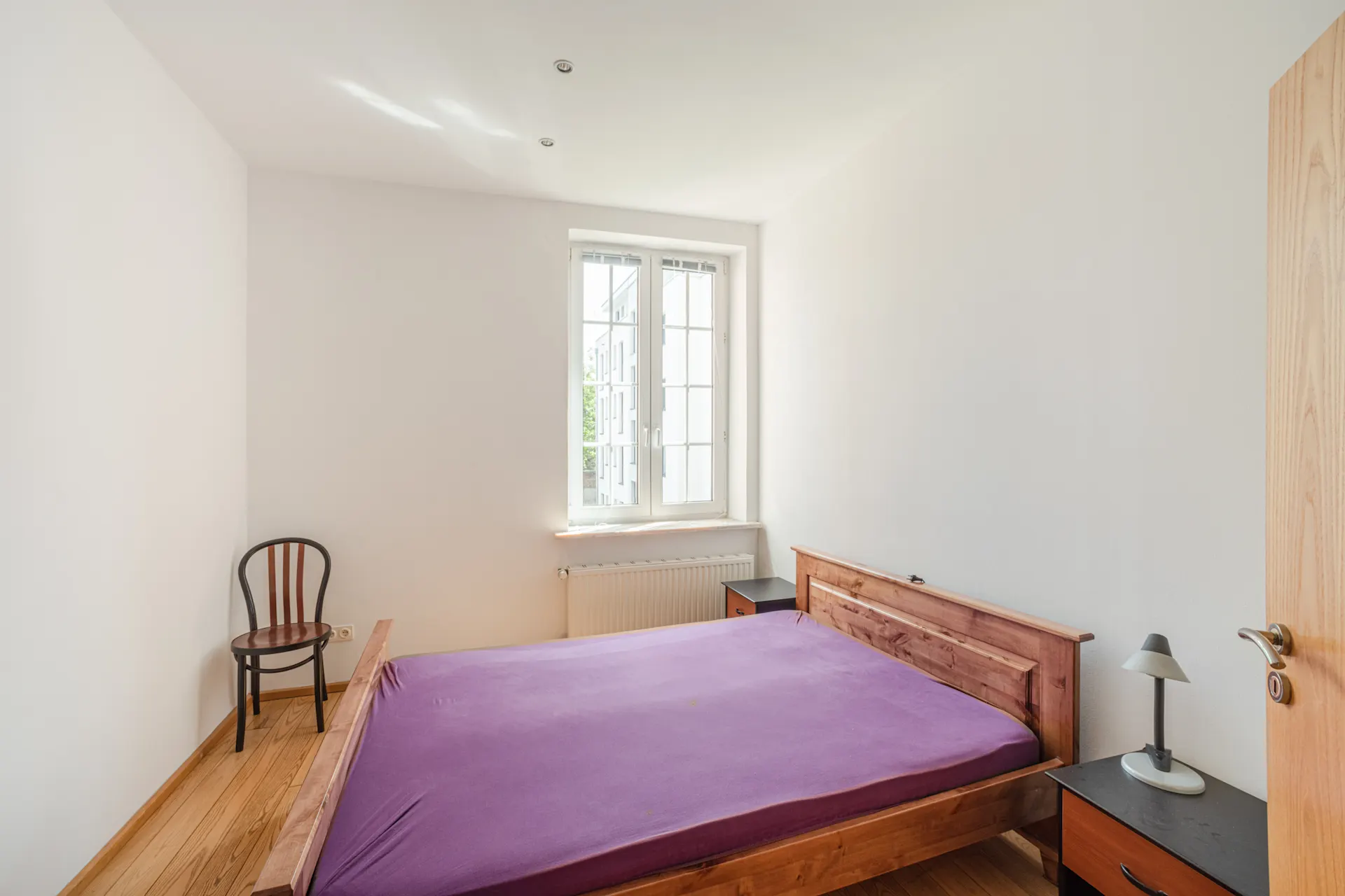 Schlafzimmer mit Holzbett, lila Bettlaken, Stuhl und Fenster, daneben zwei Nachttische.