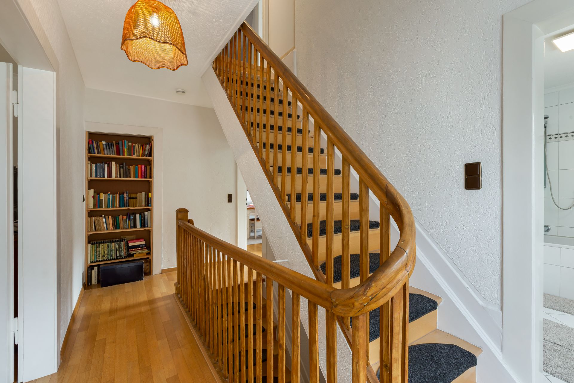 Flur mit Holztreppe, Bücherregal und Blick ins Badezimmer mit Dusche.