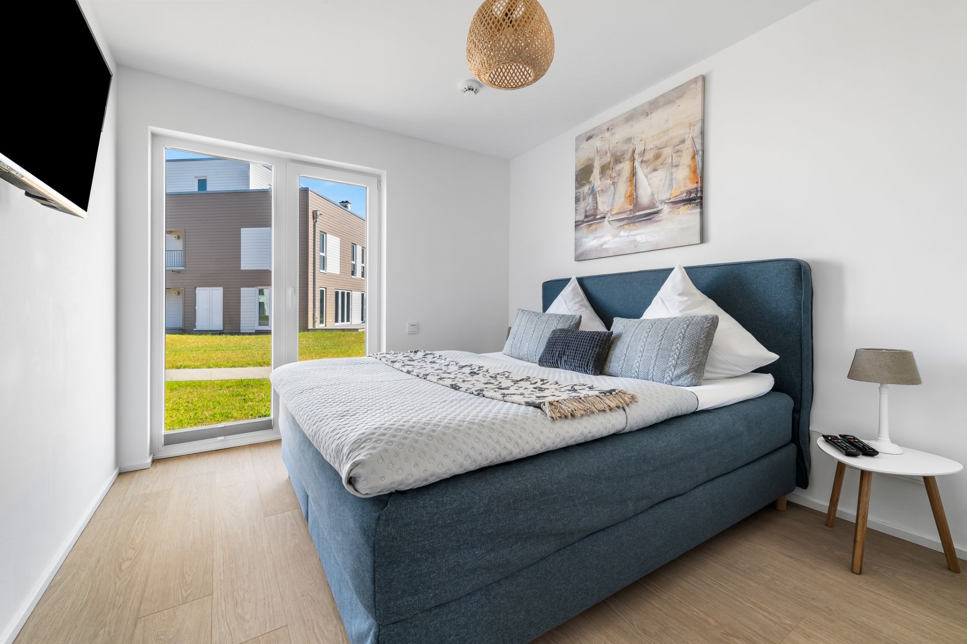 Helles Schlafzimmer mit Doppelbett, Wandbild, TV und Zugang zu einer Terrasse mit Blick auf Nachbarhäuser.