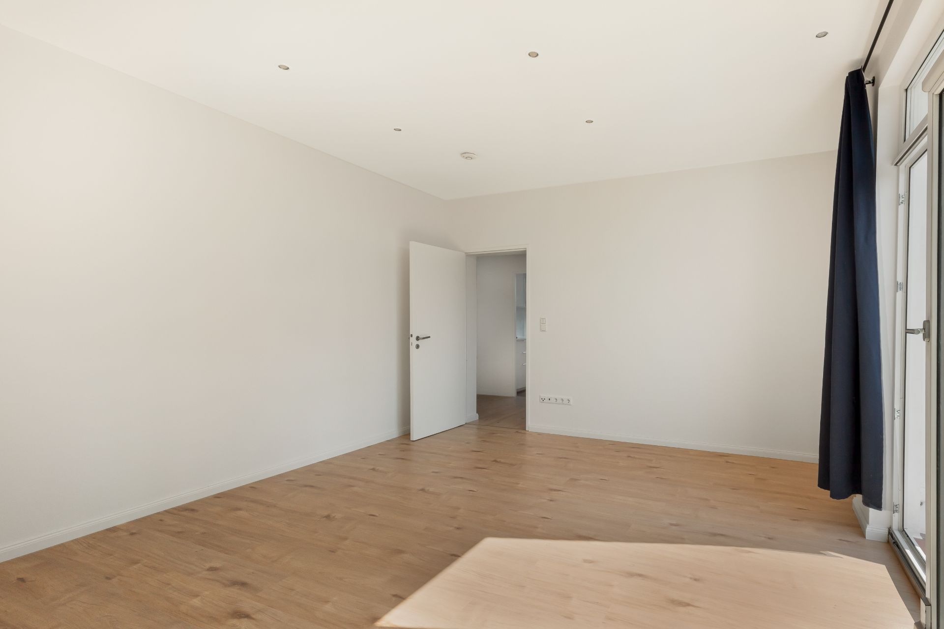 Helles, leeres Zimmer mit Holzboden, weißen Wänden und offenem Fenster mit dunklem Vorhang.