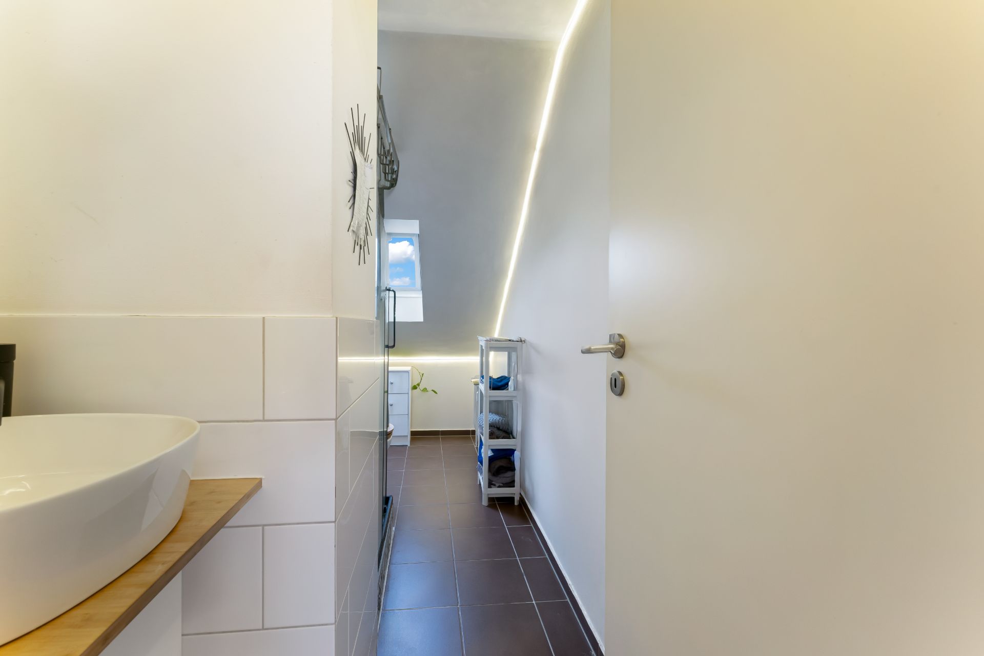 Modernes Badezimmer mit Waschbecken, geöffneter Tür und schmalem Fenster im Hintergrund.