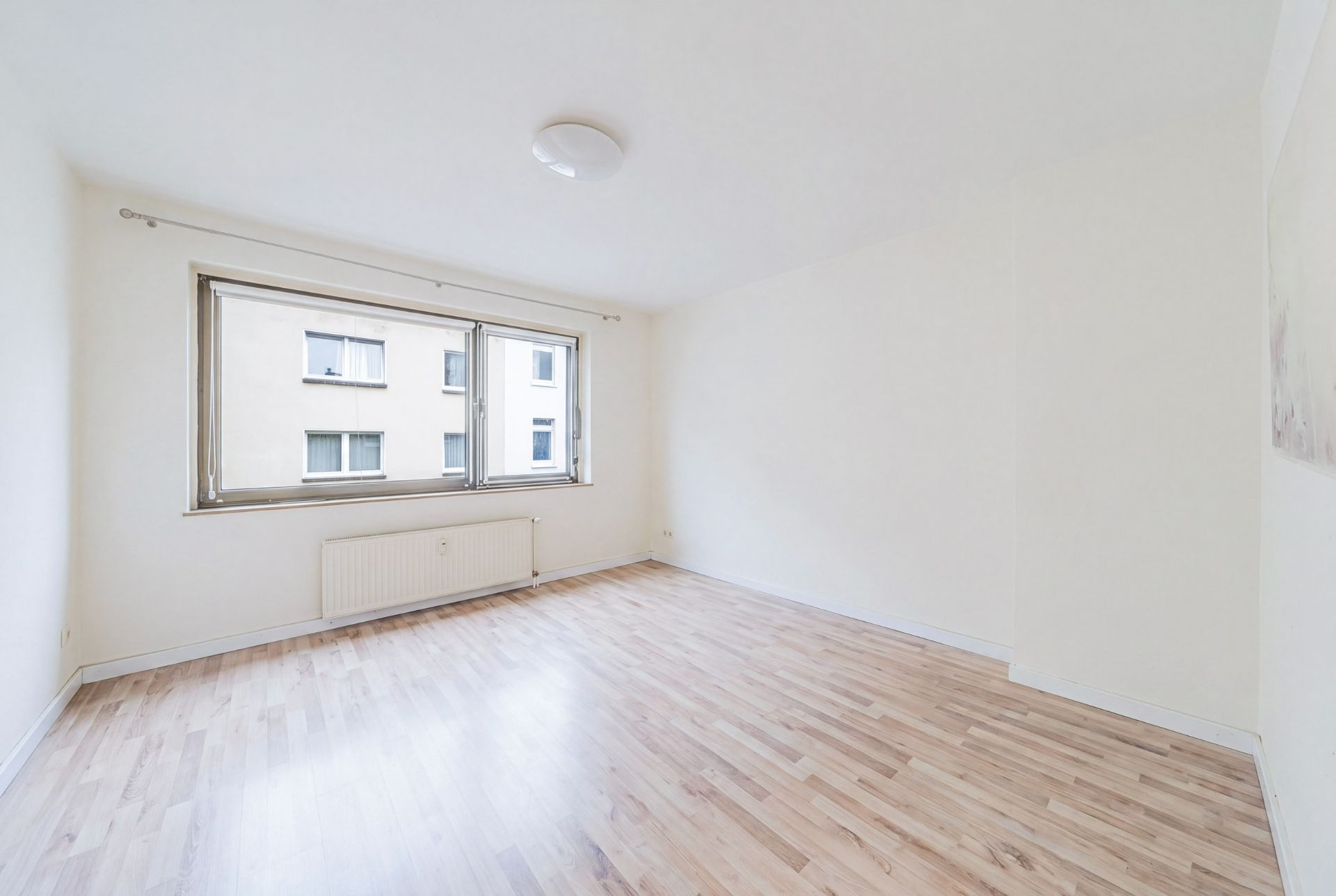 Helles, leeres Zimmer mit großem Fenster, Holzfußboden und weiß gestrichenen Wänden.