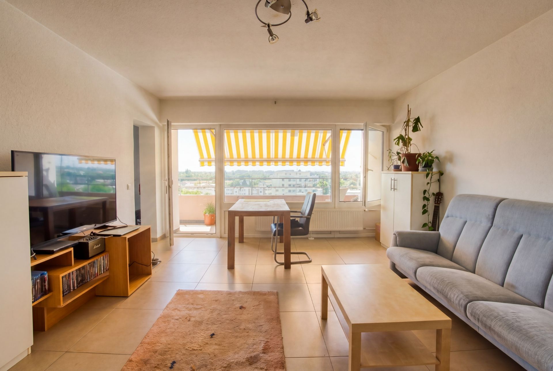 Helles Wohnzimmer mit Sofa, Fernseher, Esstisch und großem Fenster mit gelb-weißer Markise.