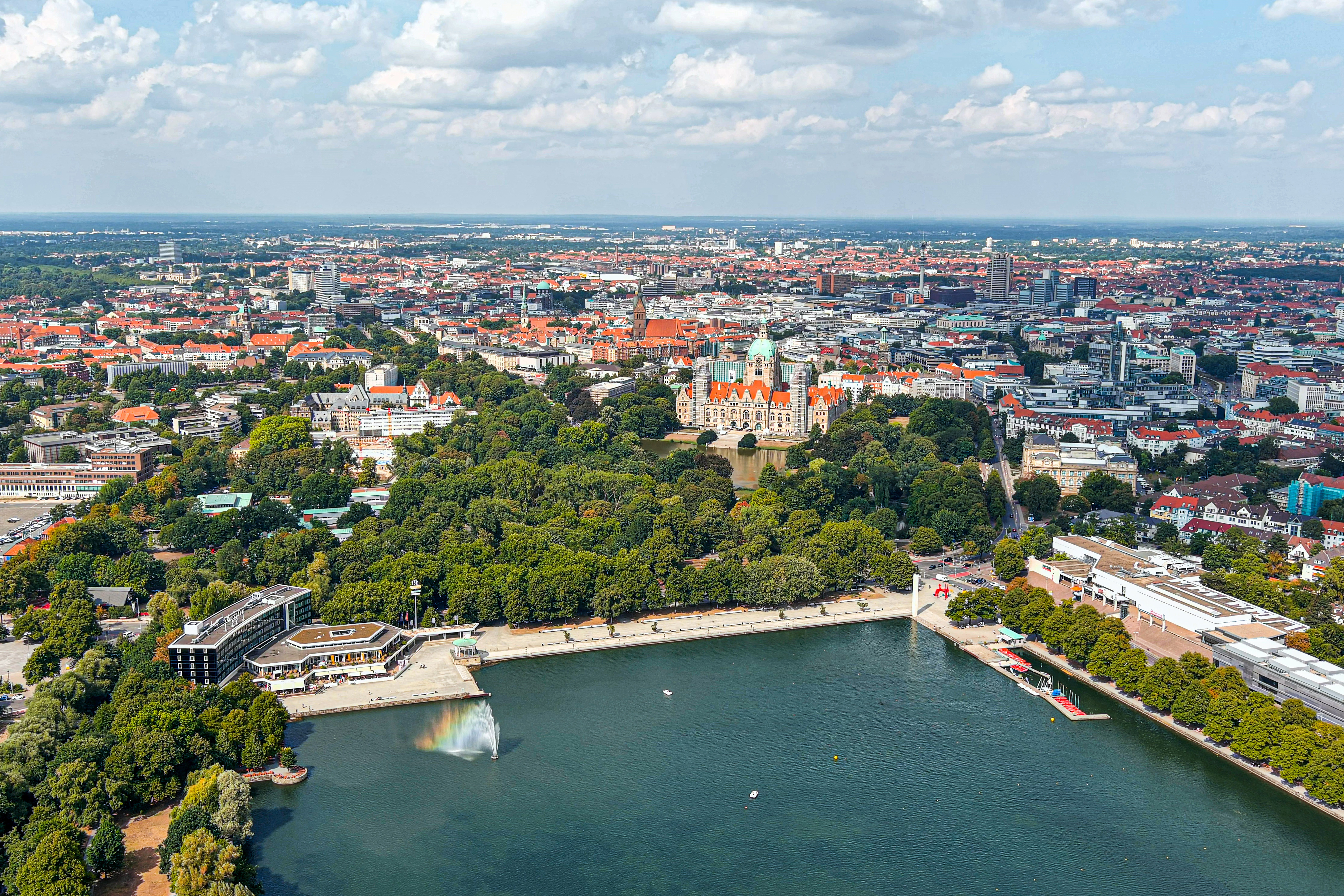 Luftaufnahme einer Stadt mit See, Springbrunnen, vielen Bäumen und historischen Gebäuden.