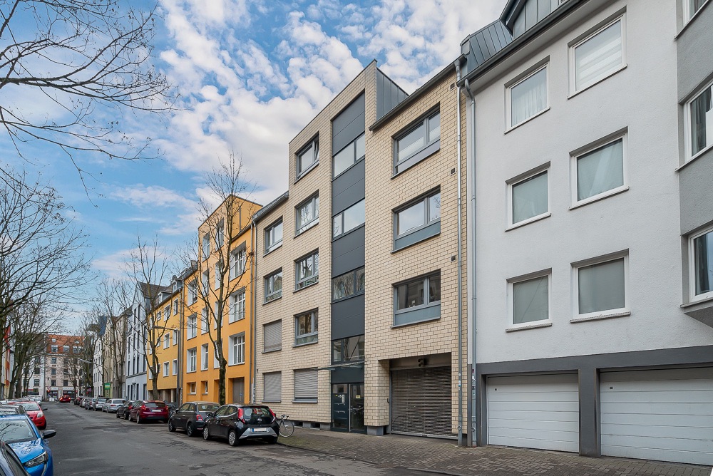 Mehrstöckiges Wohnhaus mit Parkplätzen und Autos an einer ruhigen Straße.