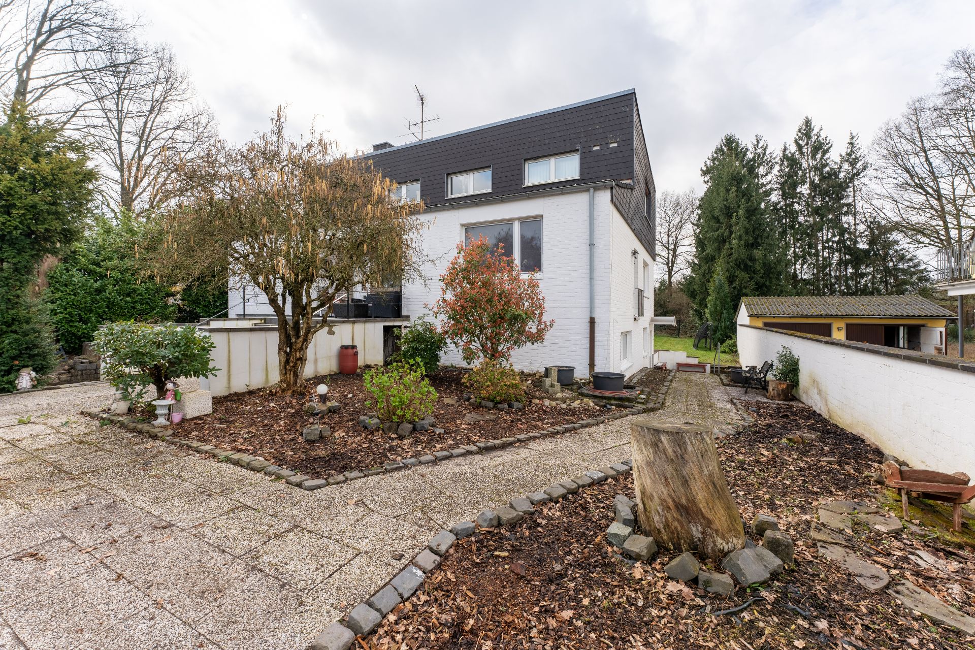 Gepflegter Garten mit Bäumen und Sträuchern vor einem weißen Wohnhaus mit dunklem Dach.