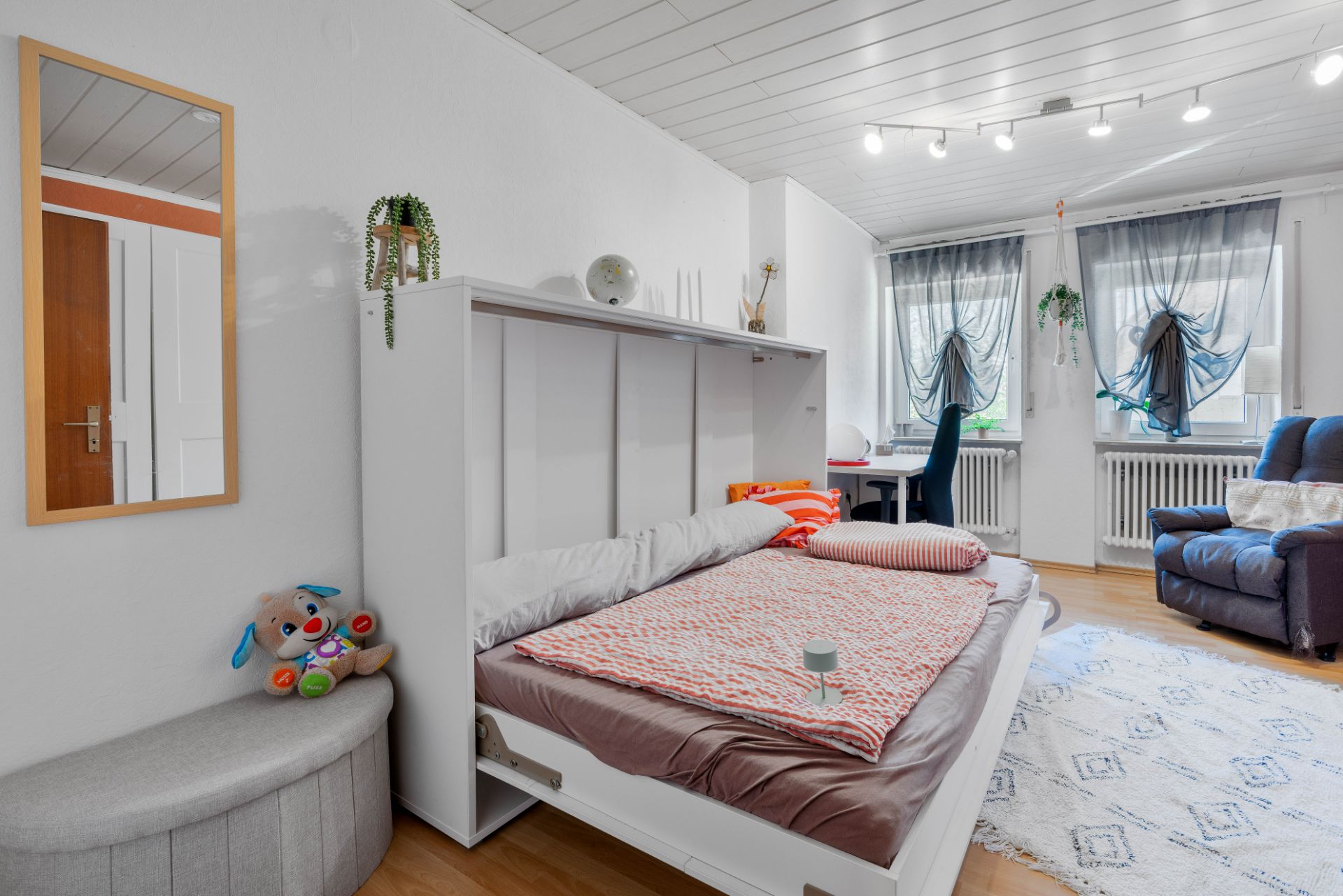 Helles Schlafzimmer mit ausgeklapptem Bett, Sessel, Schreibtisch und zwei Fenstern mit Vorhängen.