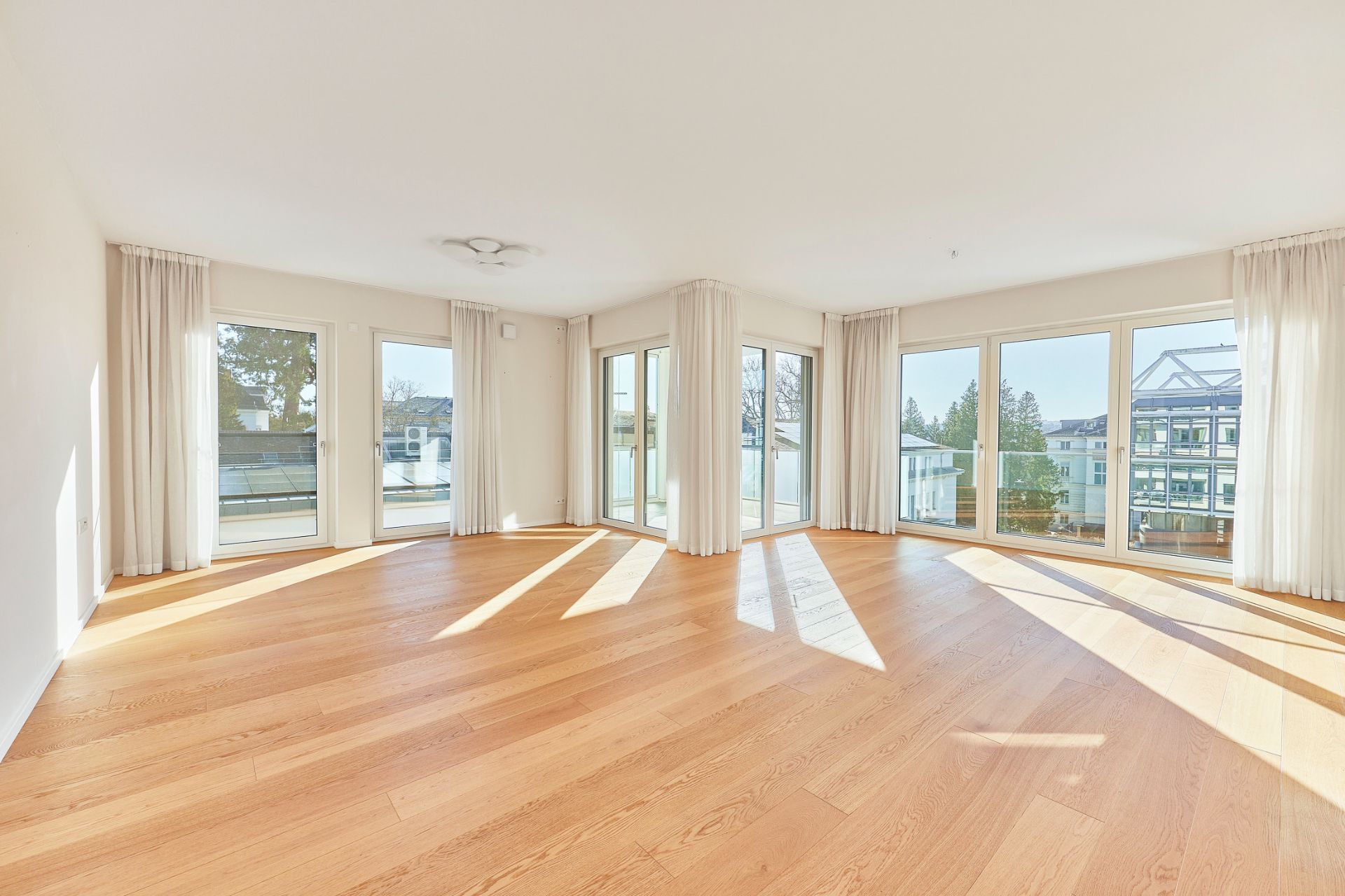 Helles, leeres Wohnzimmer mit großen Fenstern, Parkettboden und weißen Vorhängen.