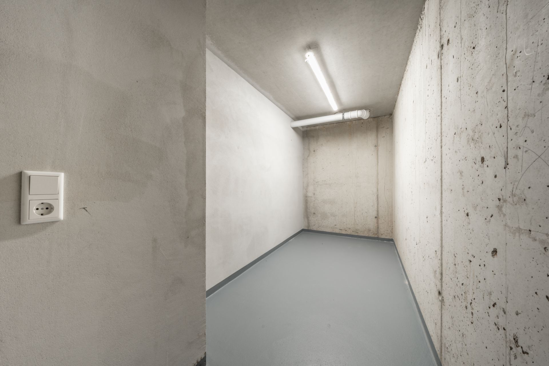 Leerer, schmaler Raum mit Betonwänden und grauem Boden, beleuchtet durch eine Deckenleuchte.