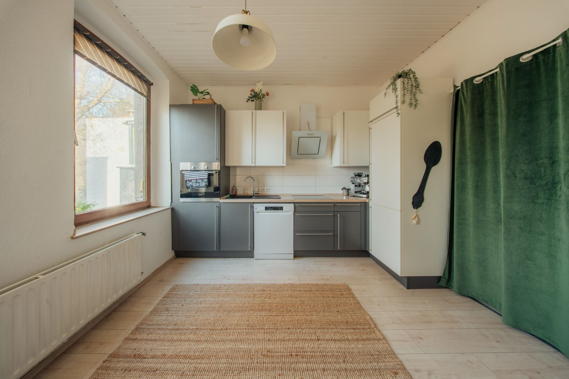 Helle Küche mit grauen und weißen Schränken, großem Fenster und grünem Vorhang.