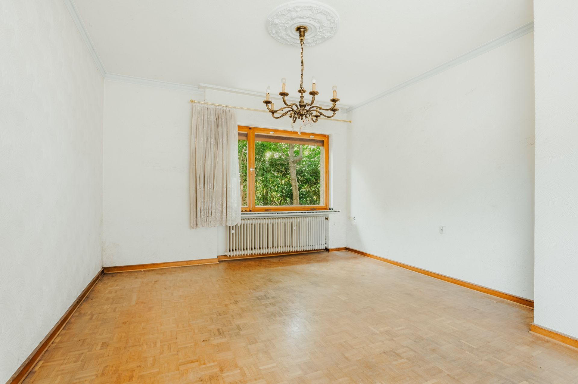 Helles, leeres Zimmer mit Parkettboden, Kronleuchter und großem Fenster mit Blick ins Grüne.