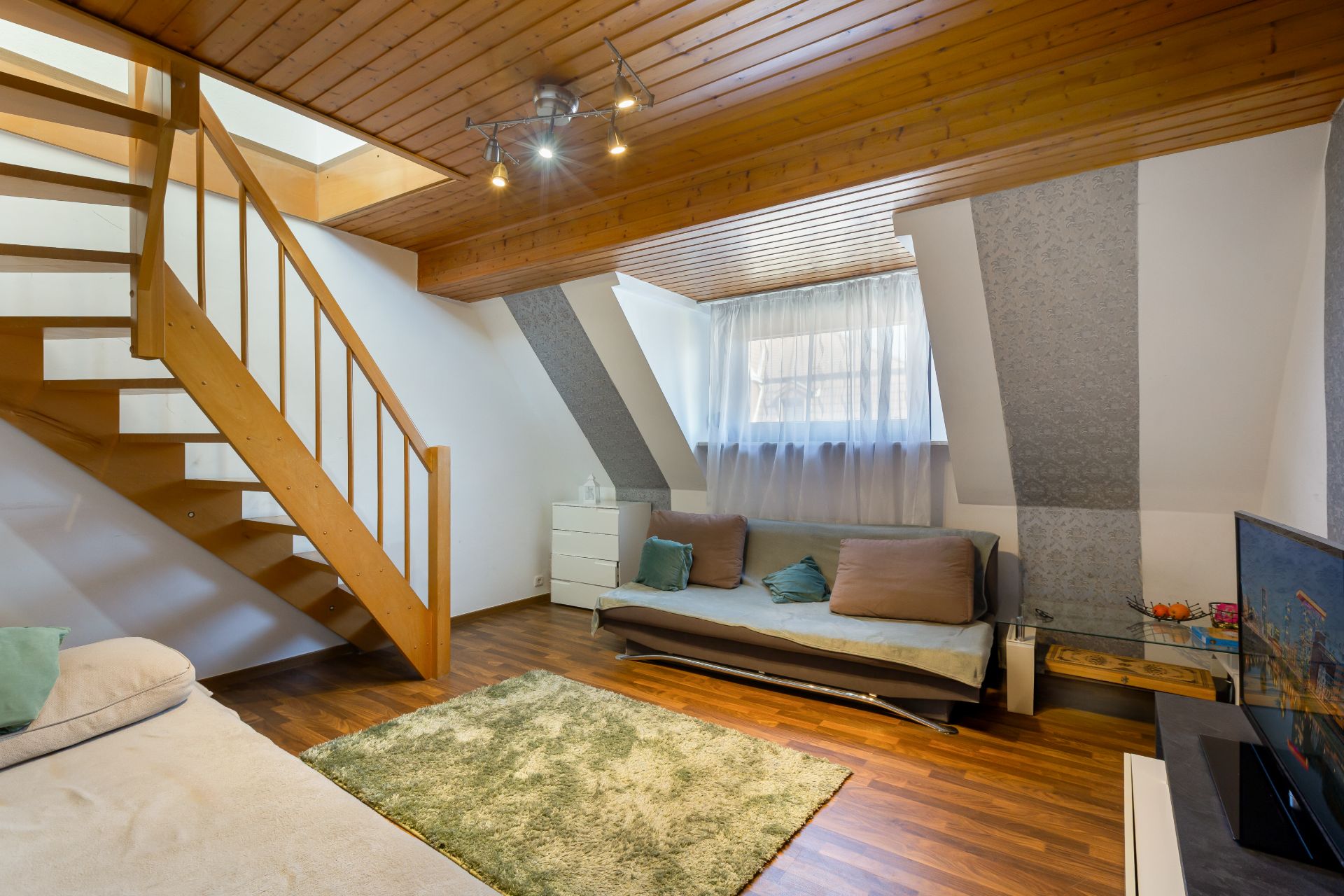 Wohnzimmer mit Holzdecke, offener Treppe, Sofa, grünem Teppich und Fernseher.