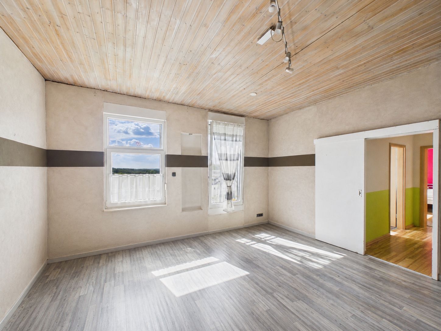 Helles, leeres Zimmer mit zwei Fenstern, hellem Holzboden und Holzdecke.