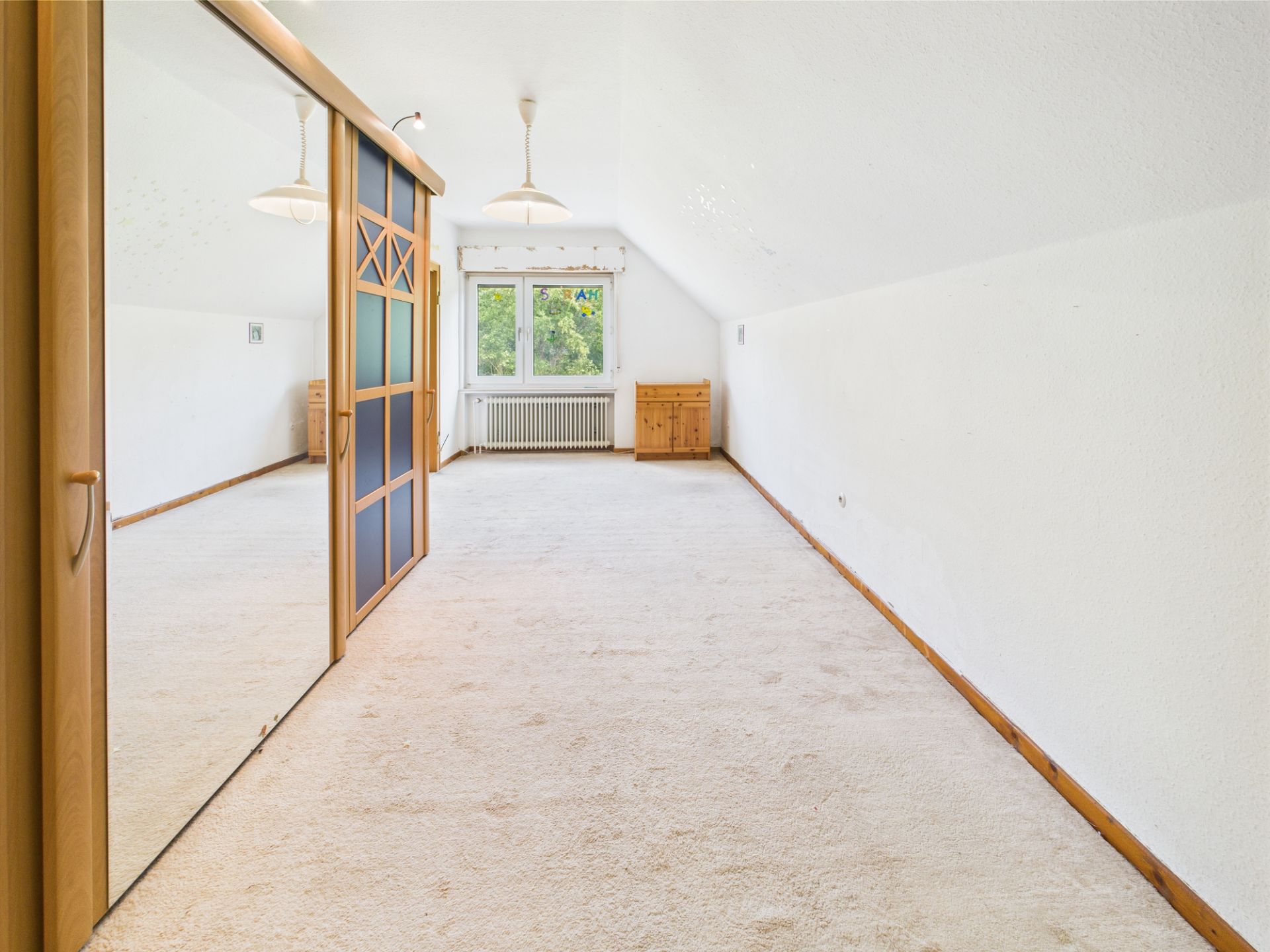 Langer, heller Raum mit Teppichboden, Einbauschrank mit Spiegel und Fenster am Ende.