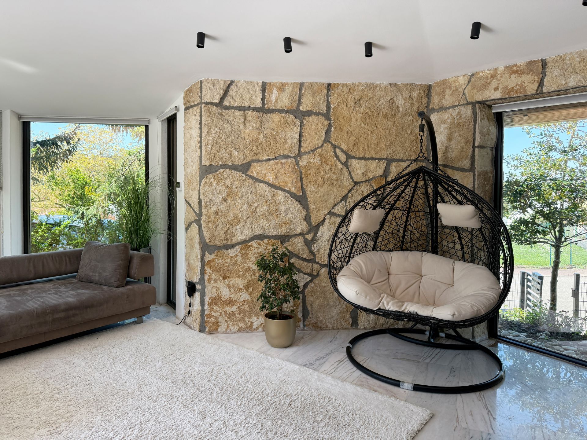 Wohnzimmer mit Steinwand, Hängesessel, braunem Sofa, Pflanze und großen Fenstern mit Gartenblick.