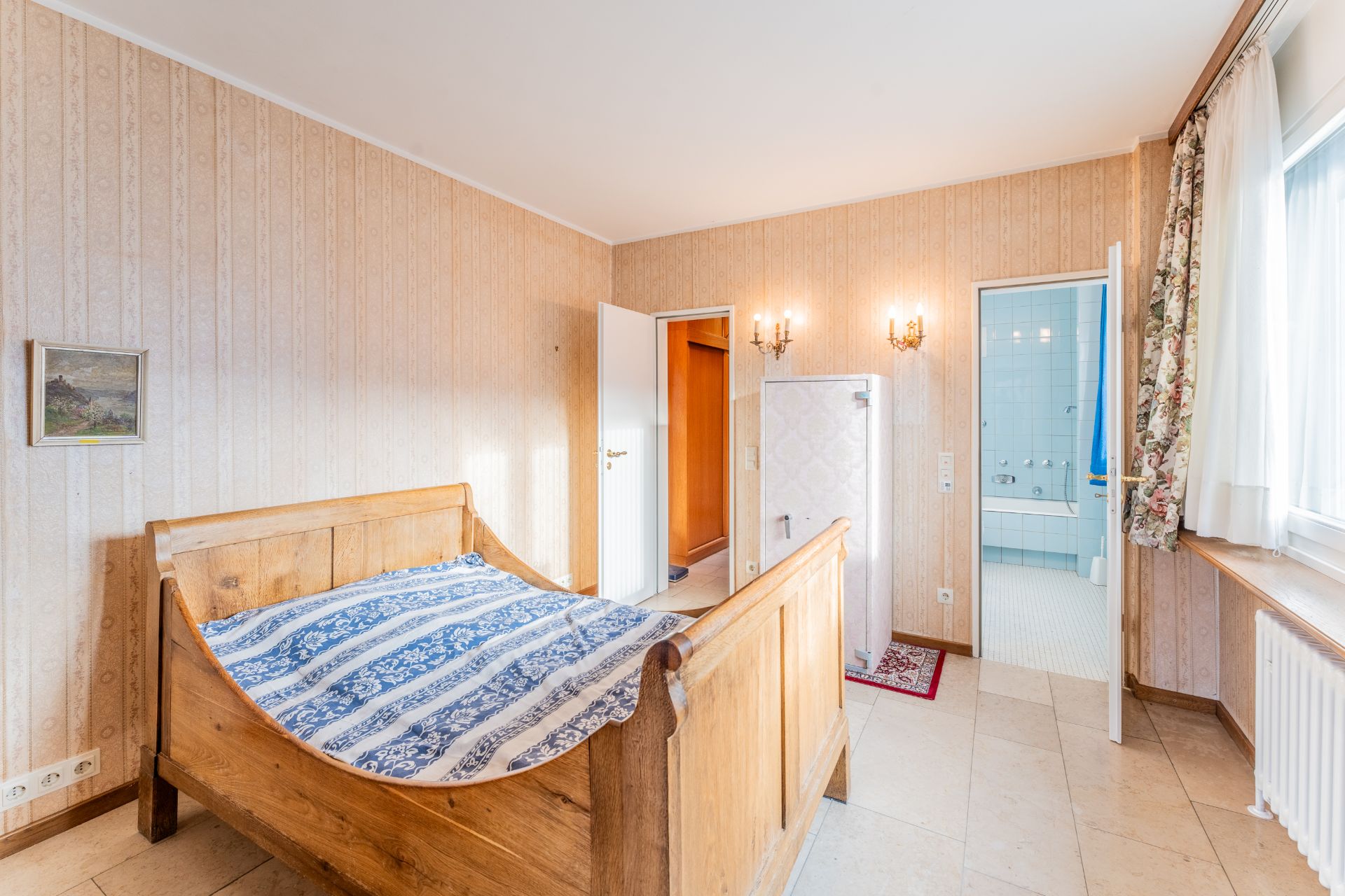 Helles Schlafzimmer mit Holzbett, gemusterter Decke und Zugang zu einem Badezimmer mit blauen Fliesen.