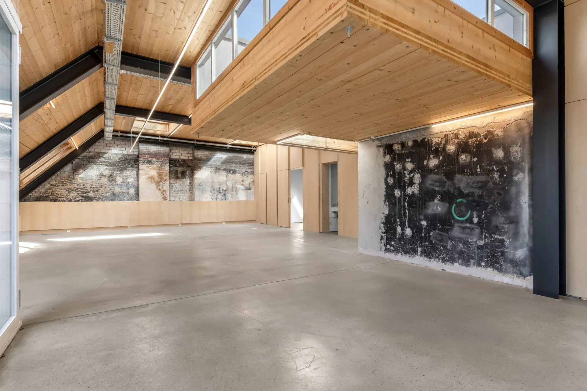 Großer, heller Raum mit Holzdecke, Betonboden und einer Wand mit sichtbarem Mauerwerk.