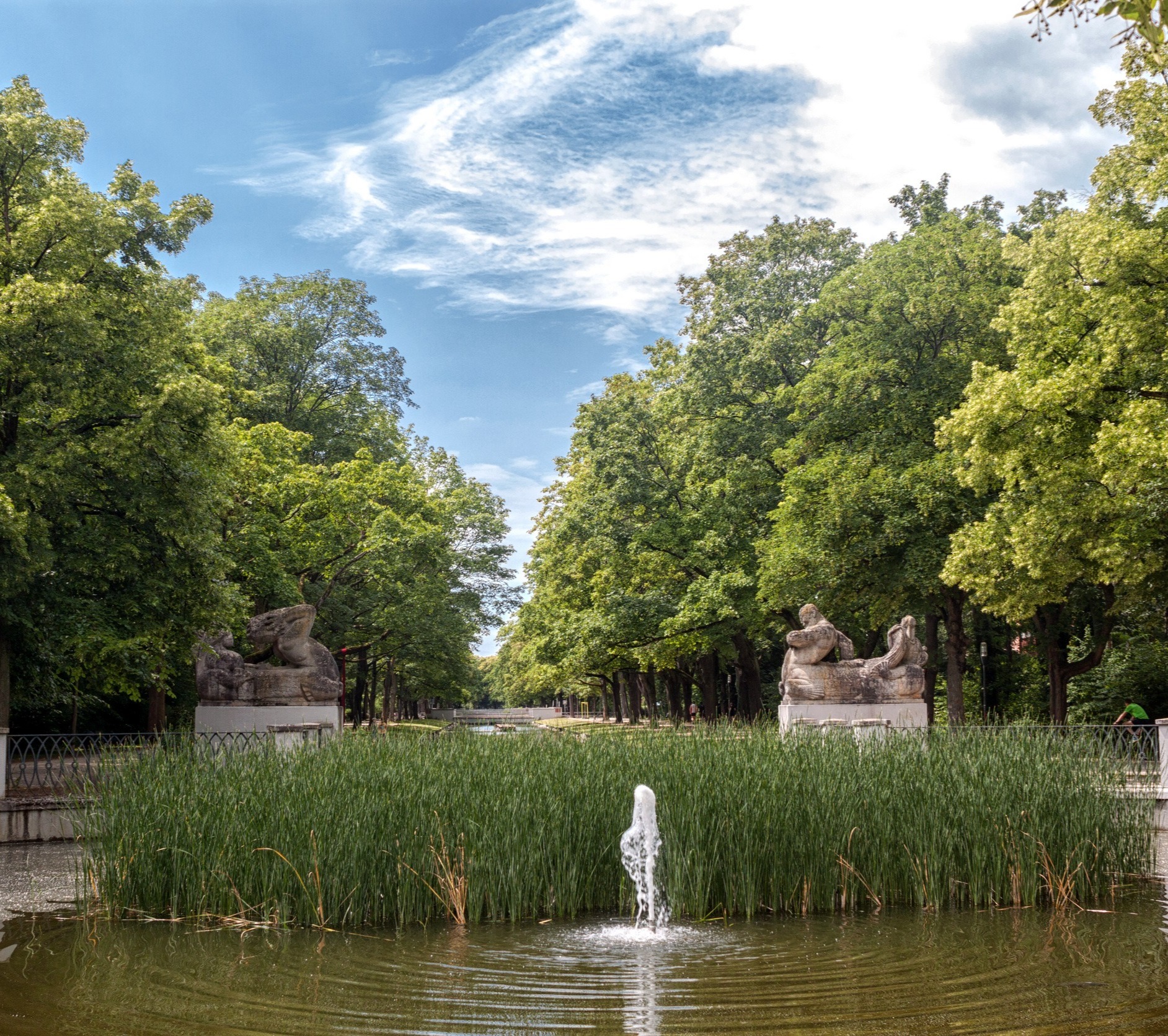 Brunnen mit Wasserstrahl, umgeben von Schilf und Bäumen in einer Parkanlage.