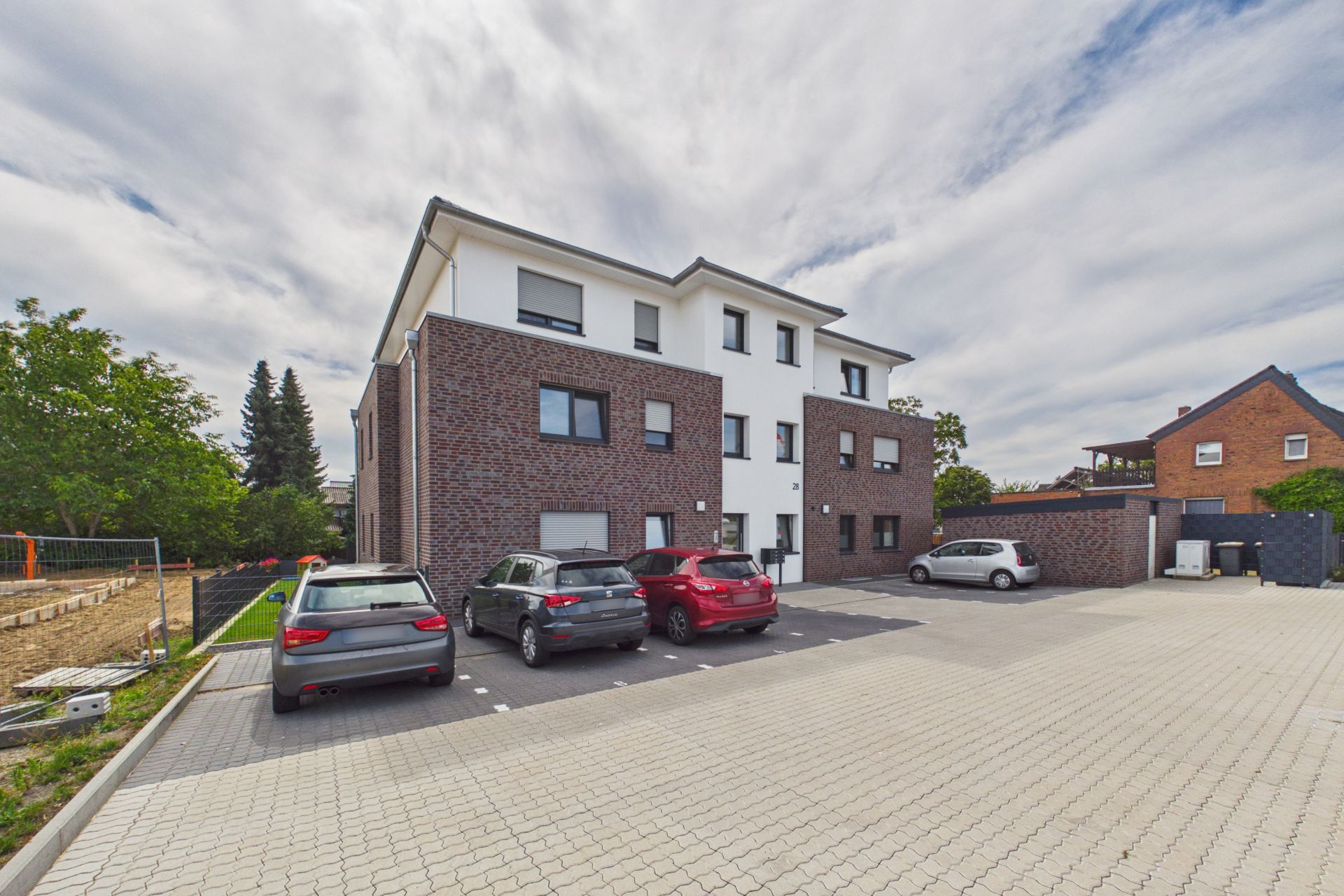 Mehrfamilienhaus mit großen Fenstern und Parkplatz mit mehreren Autos davor.